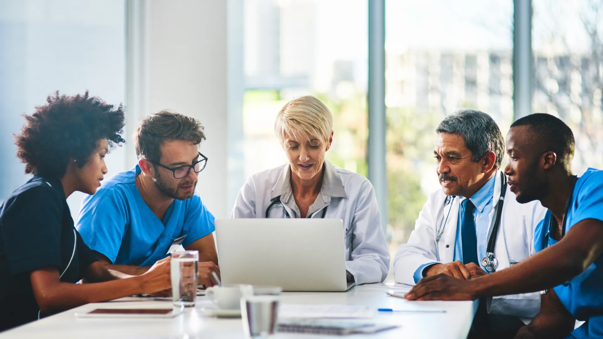Medical team in scrubs and lab coats collaborating around a laptop in a bright office meeting room.