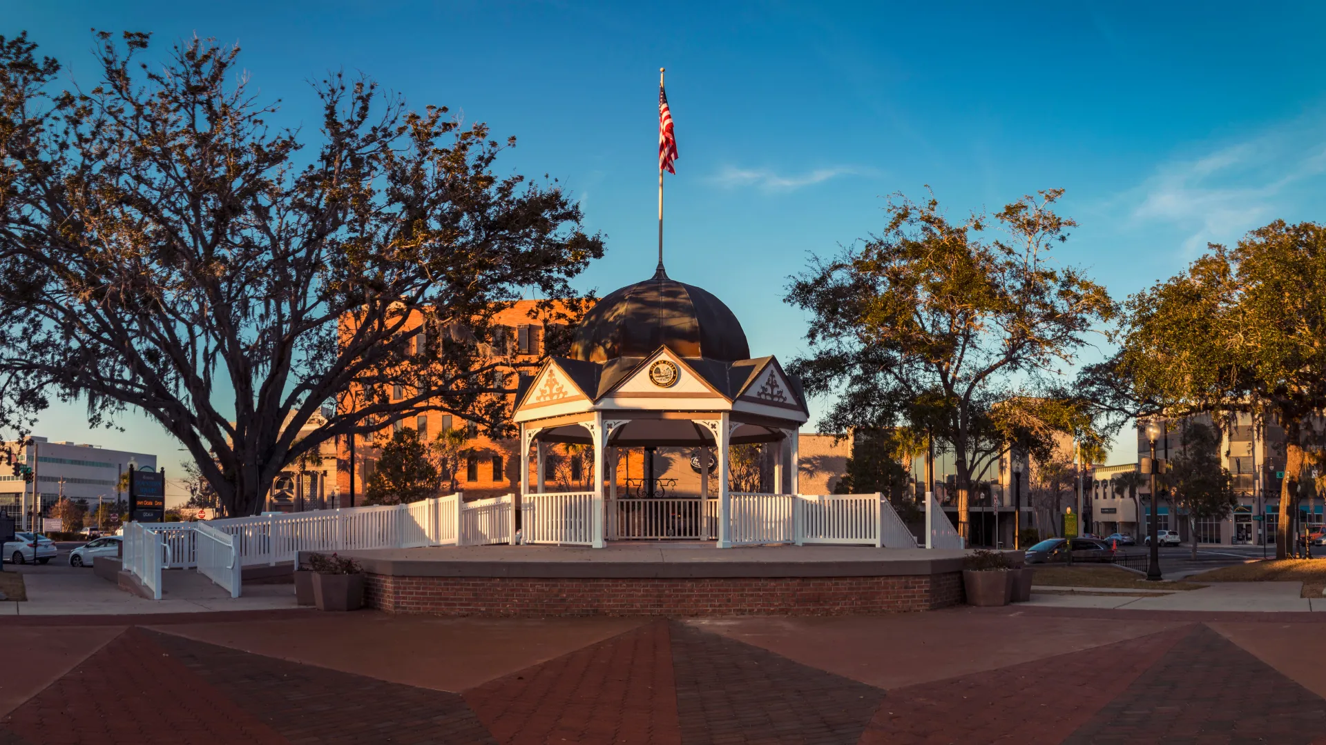 Historic white gazebo with a black dome and American flag in a sunlit town square surrounded by trees and buildings.