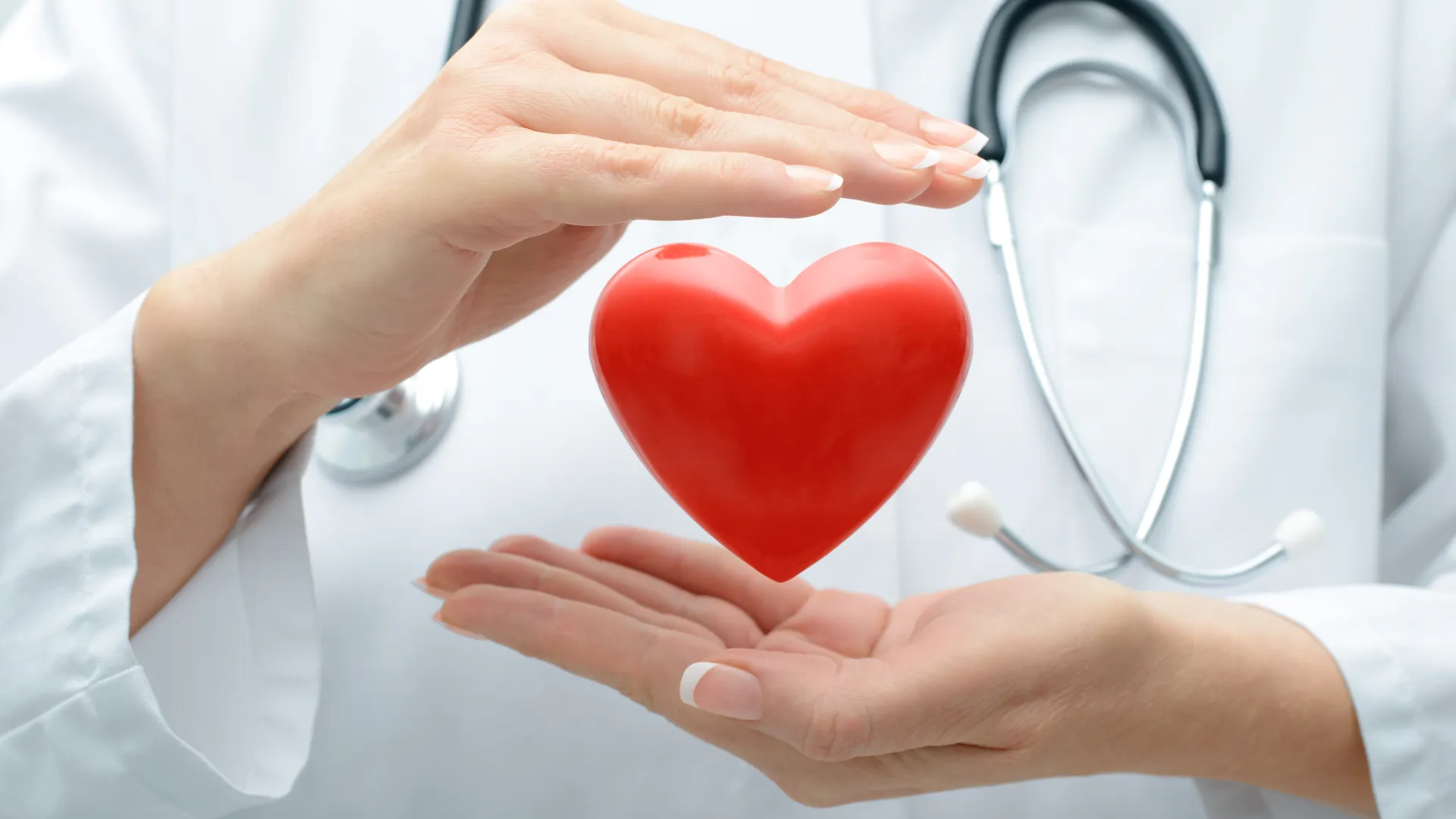Doctor in white coat with stethoscope holding a red heart symbol between hands representing heart care.
