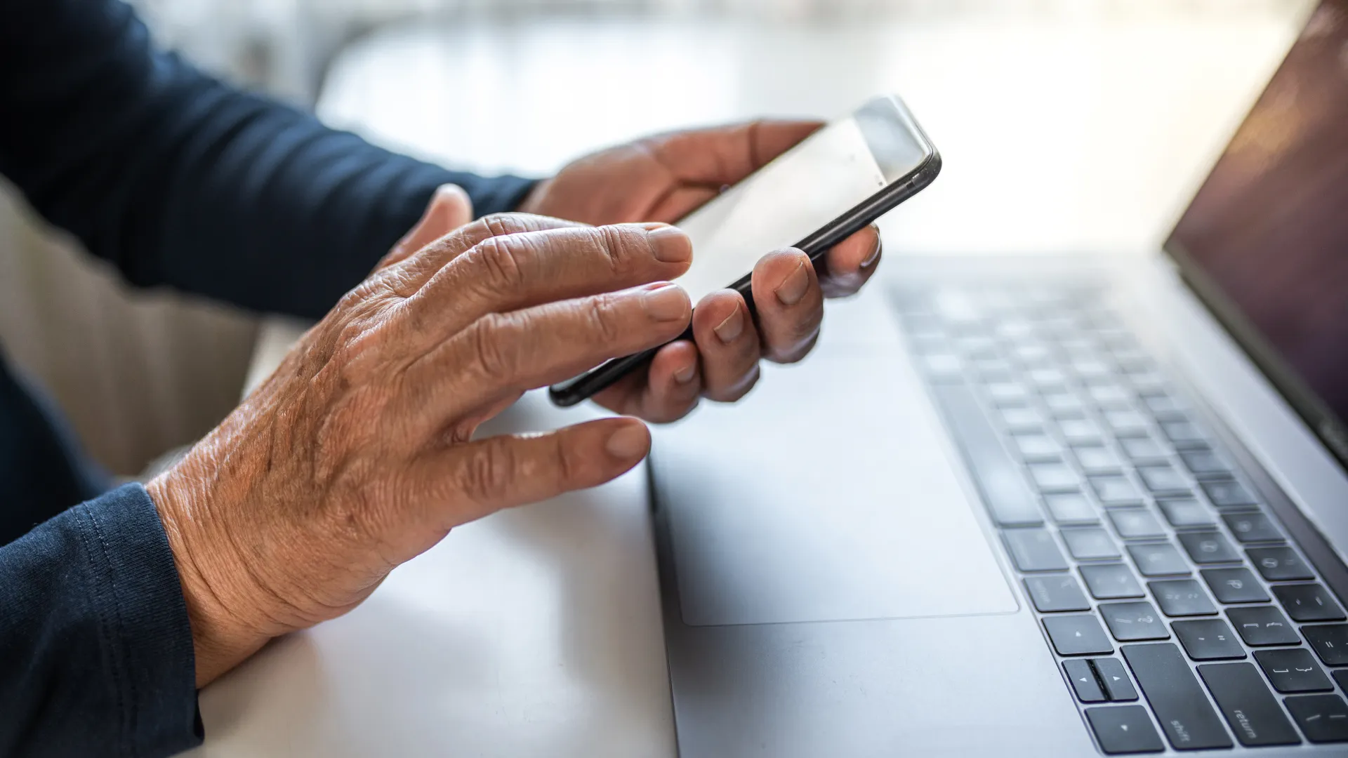 Senior person using smartphone next to open laptop on desk with natural light background.