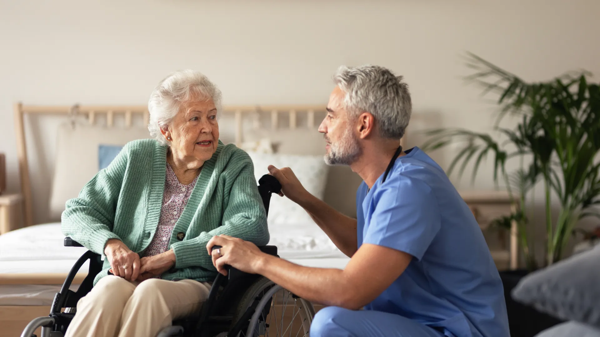 Elderly woman in a wheelchair talking to a male healthcare professional in a home setting.