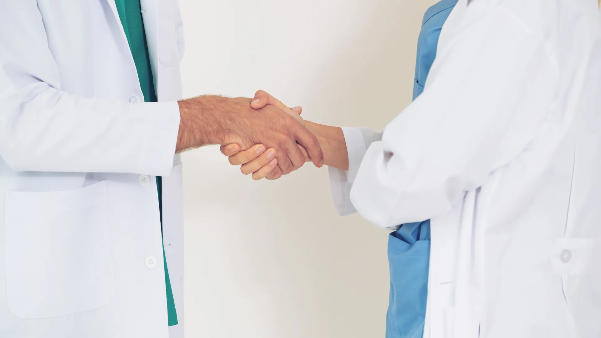 Two healthcare professionals in white coats shaking hands in a clinical setting.