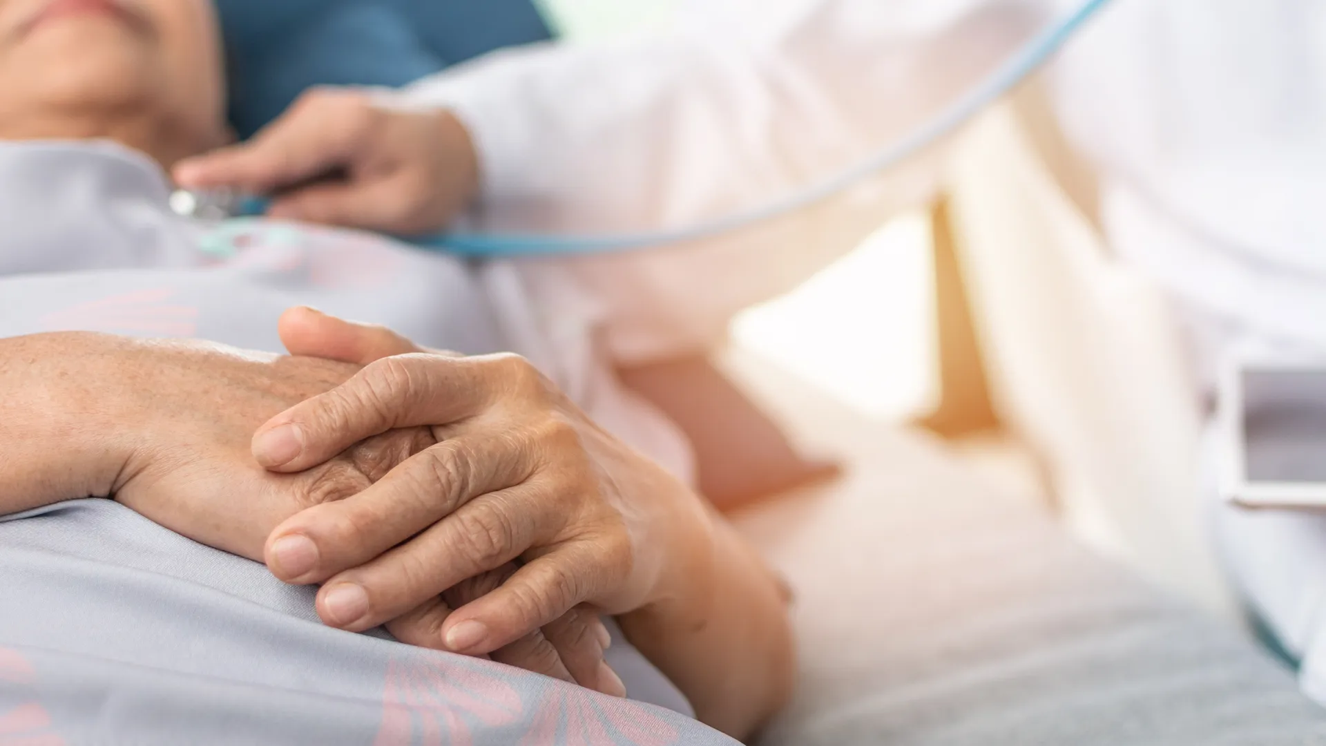 Doctor holding senior patient's hand while checking heartbeat with stethoscope, showing care and support.