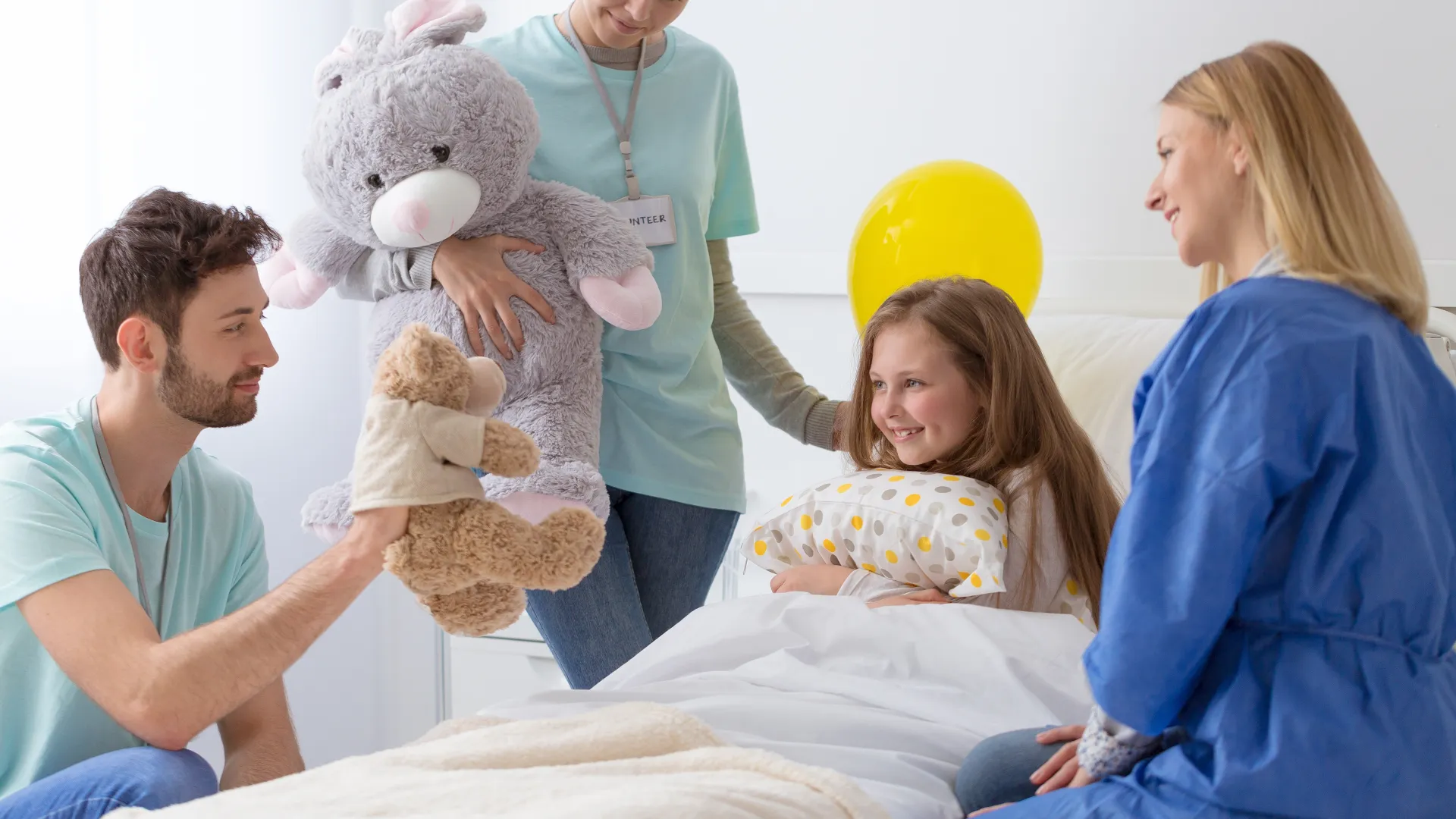 Healthcare professionals bring joy to a young girl with stuffed animals in a bright hospital room.