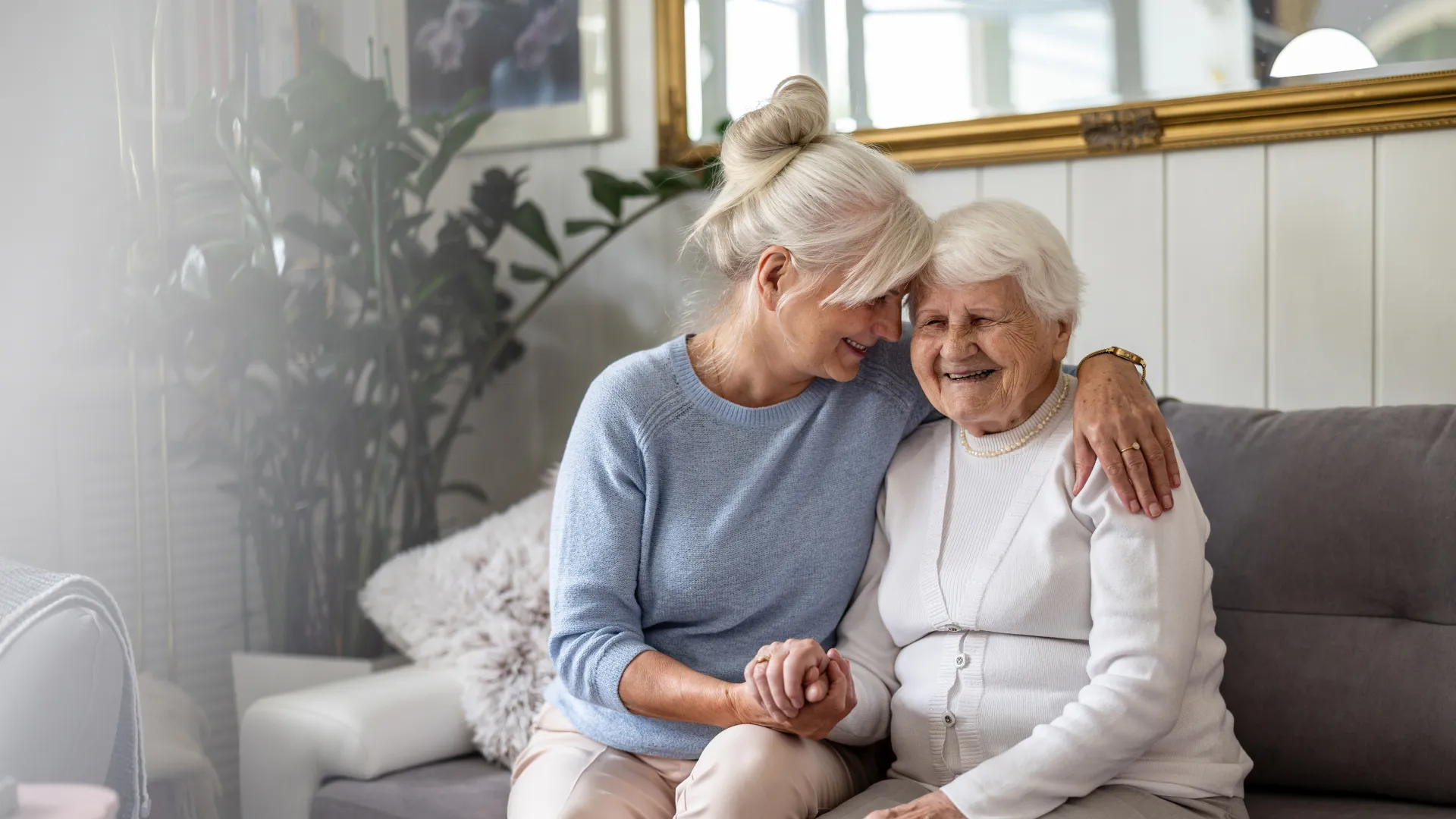 Senior woman and elderly mother sitting on sofa, smiling and holding hands in cozy living room setting.