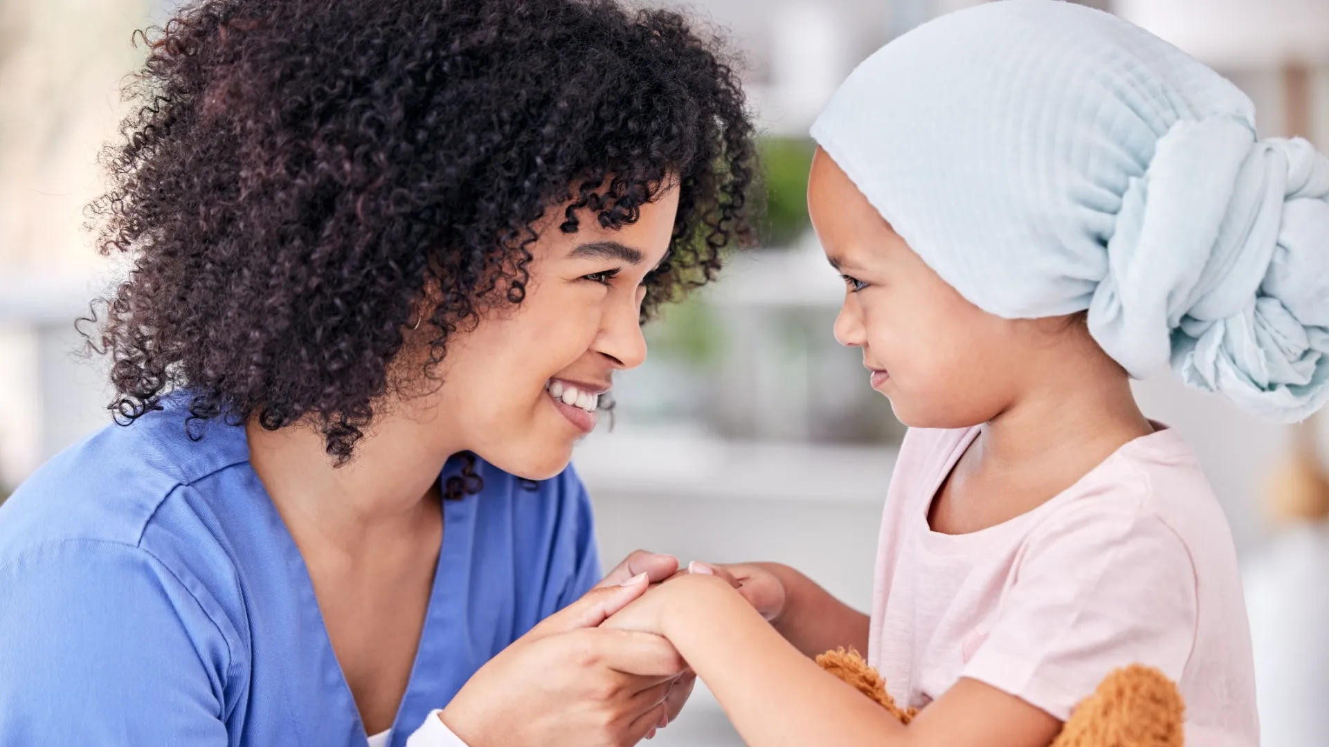 Smiling woman in blue holds hands with young child wearing headscarf and holding teddy bear, sharing a tender moment.