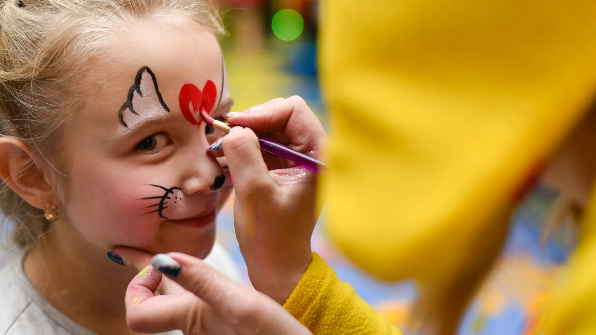 Young girl getting a colorful face painting with a cat design and red heart on her forehead