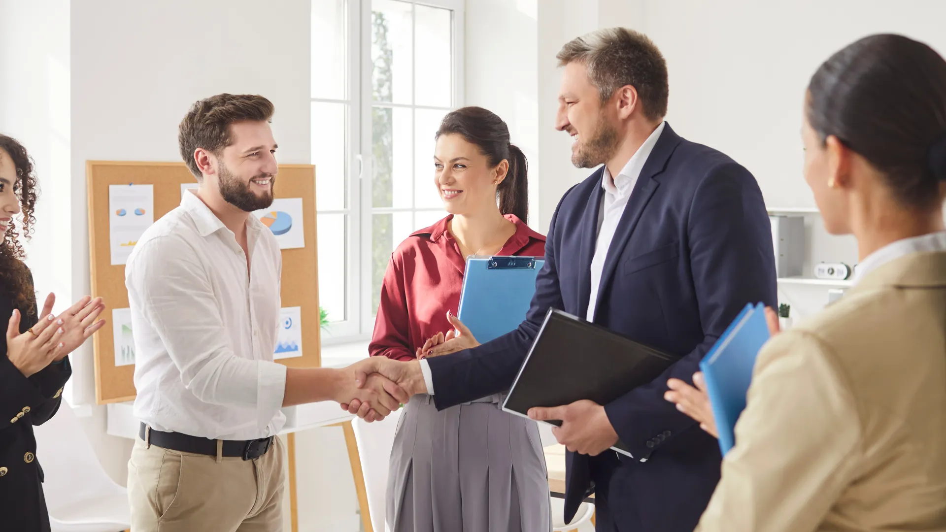 Business professionals shaking hands in a bright office while colleagues applaud during a meeting.