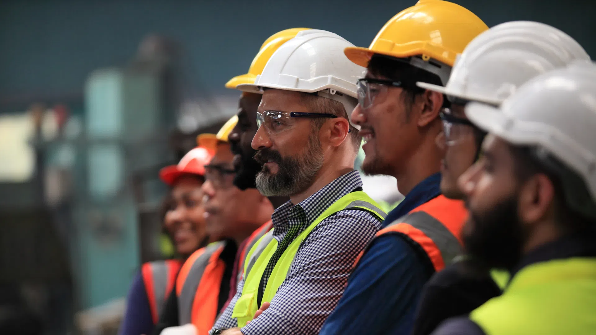 Diverse group of factory workers wearing safety helmets and reflective vests standing side by side.