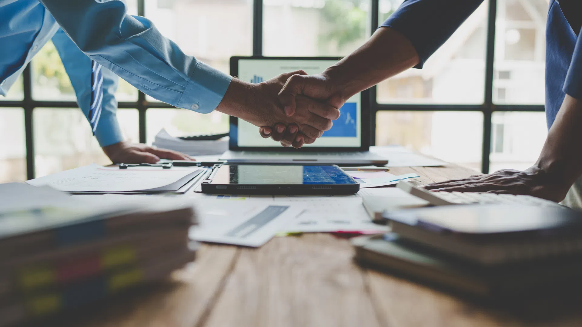 Two Essayon professionals shaking hands over a desk filled with documents and a tablet in a bright office setting