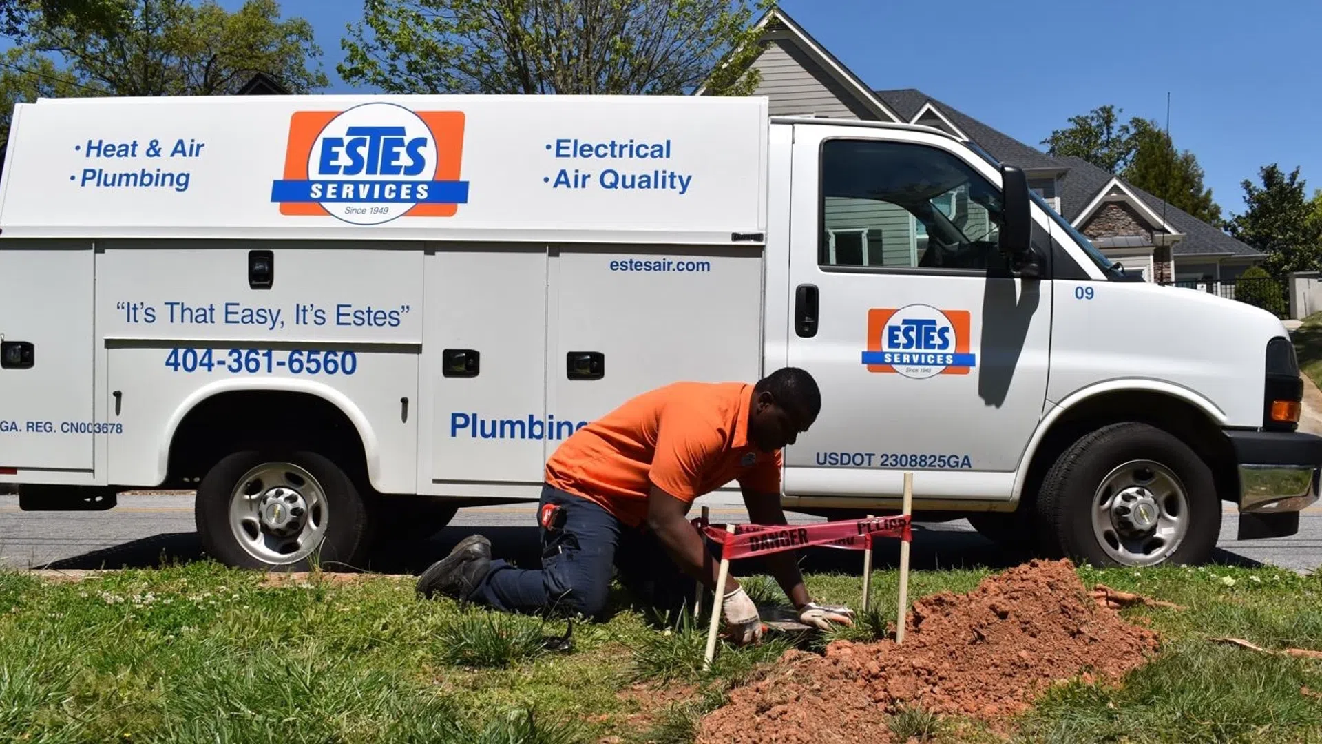 Technician in orange shirt working on plumbing near Estes Services van in suburban neighborhood.