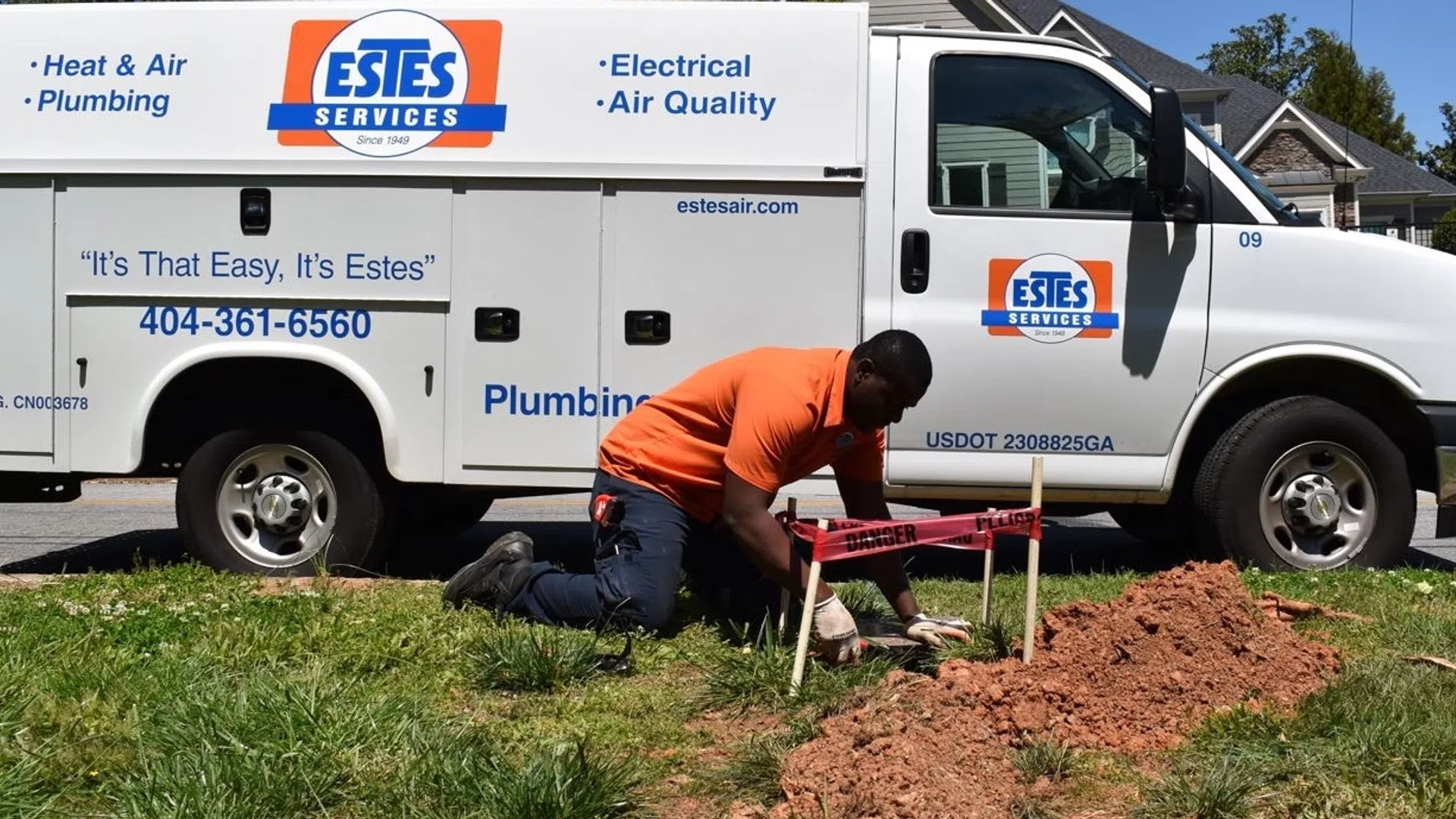 Technician in orange shirt working on plumbing repairs near Estes Services van in residential area.