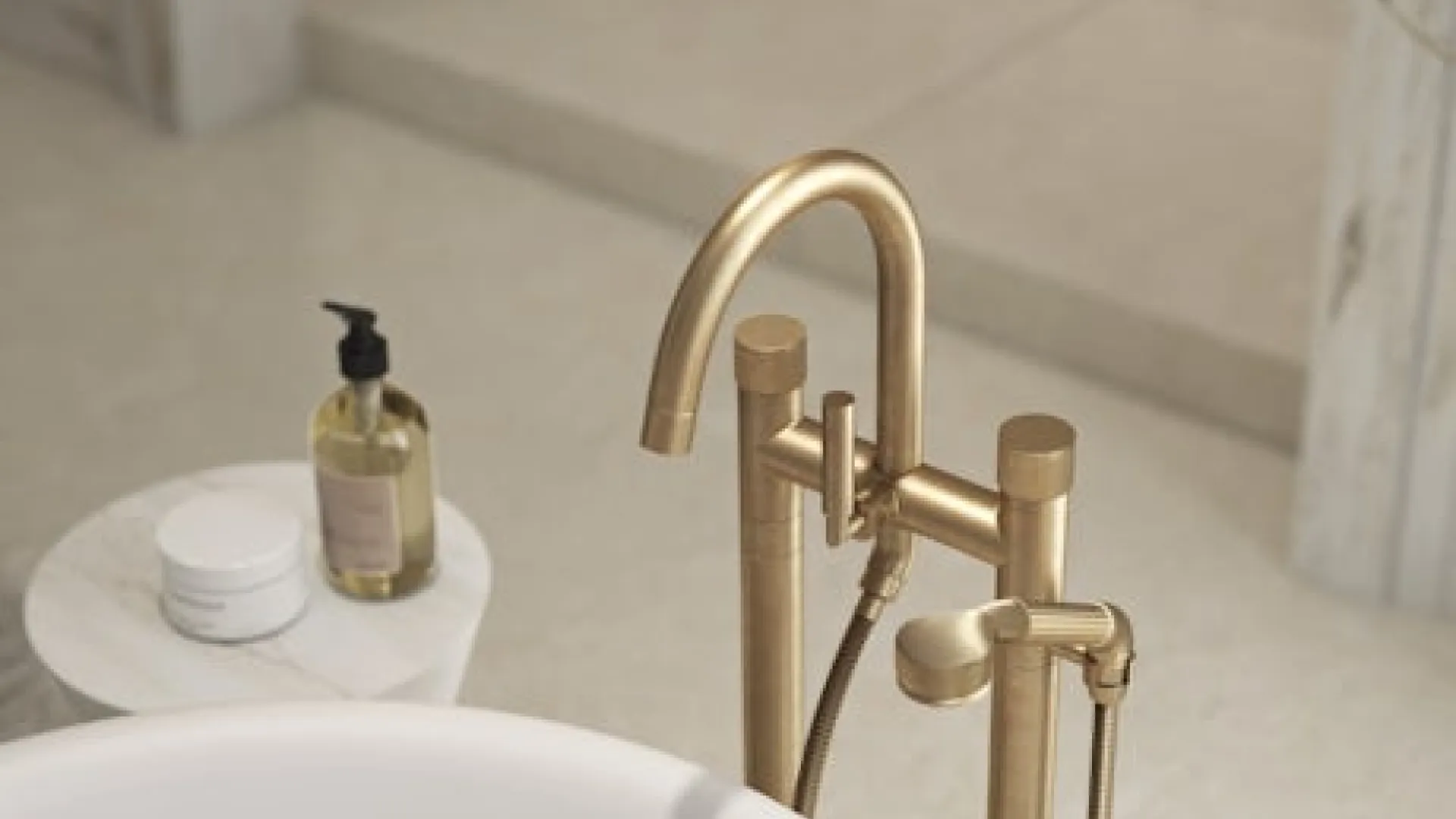 Modern gold freestanding bathtub faucet with hand shower next to a white tub in a minimalist bathroom.