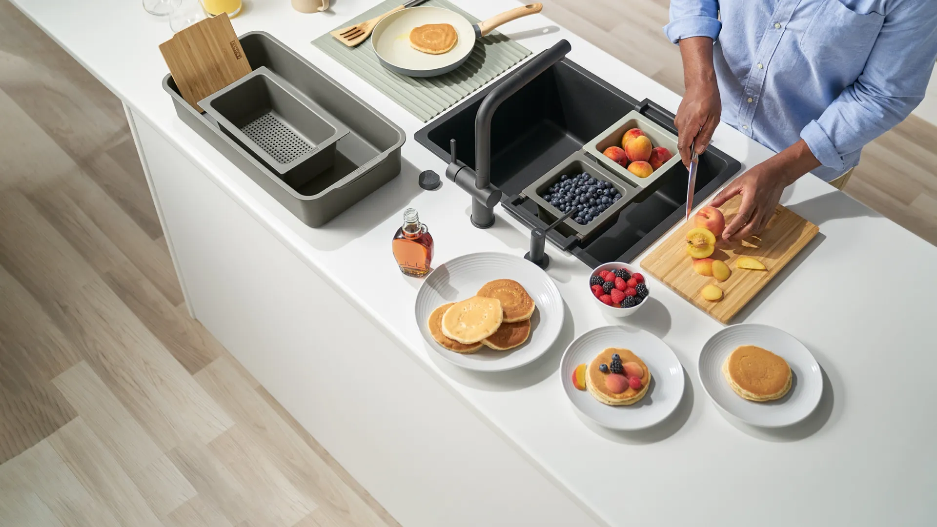 Person preparing pancakes and slicing peaches on a white kitchen island with fruits and kitchen tools.