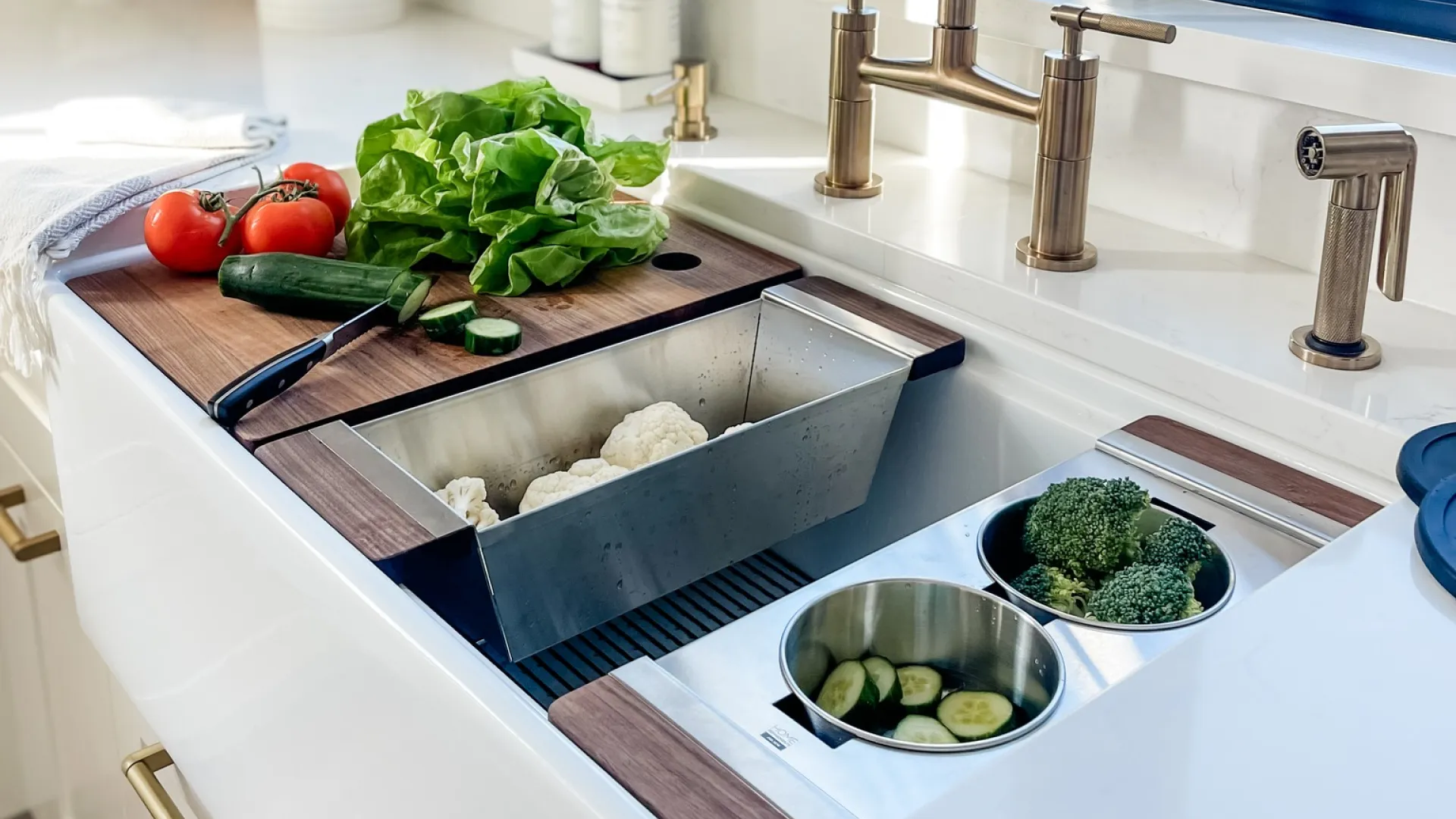Modern white kitchen sink with brass faucet, fresh vegetables, cutting board, and blue window frame in daylight.
