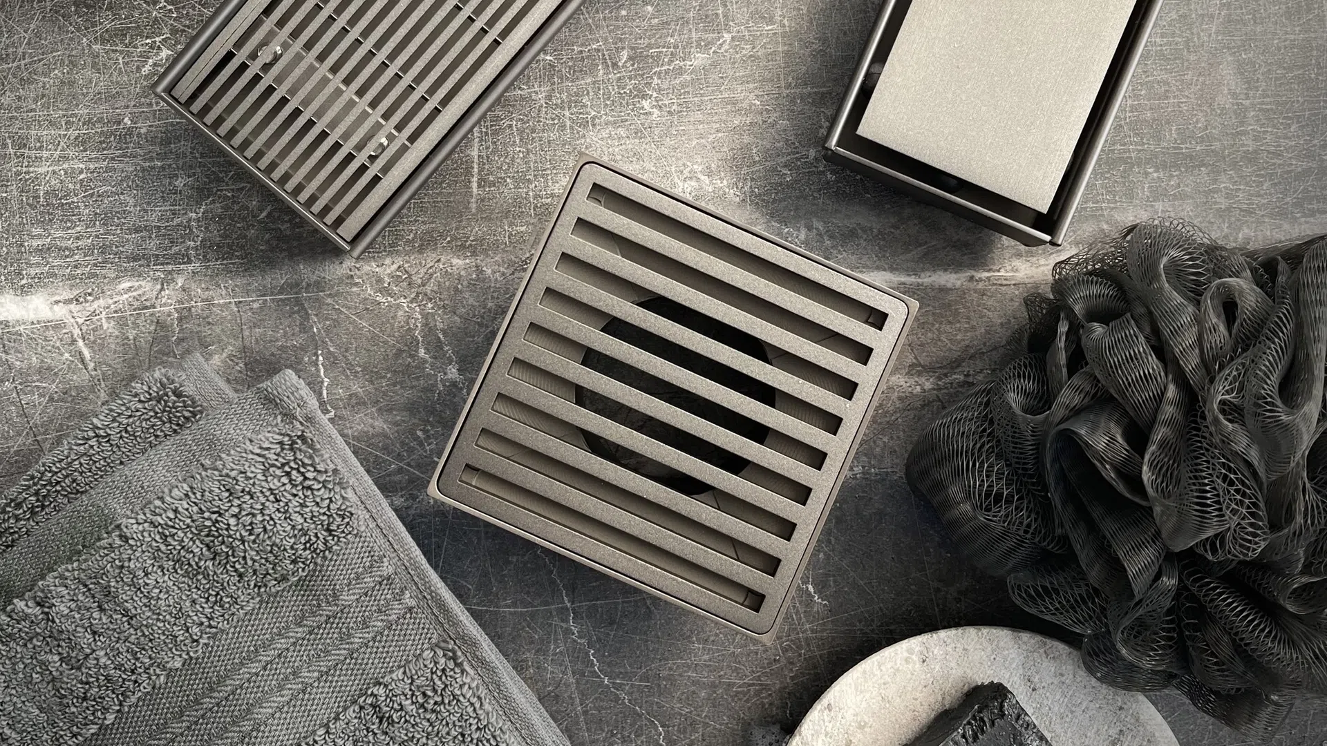 Modern metal shower drains with gray towel, soap, and dark bath sponge on textured gray surface.