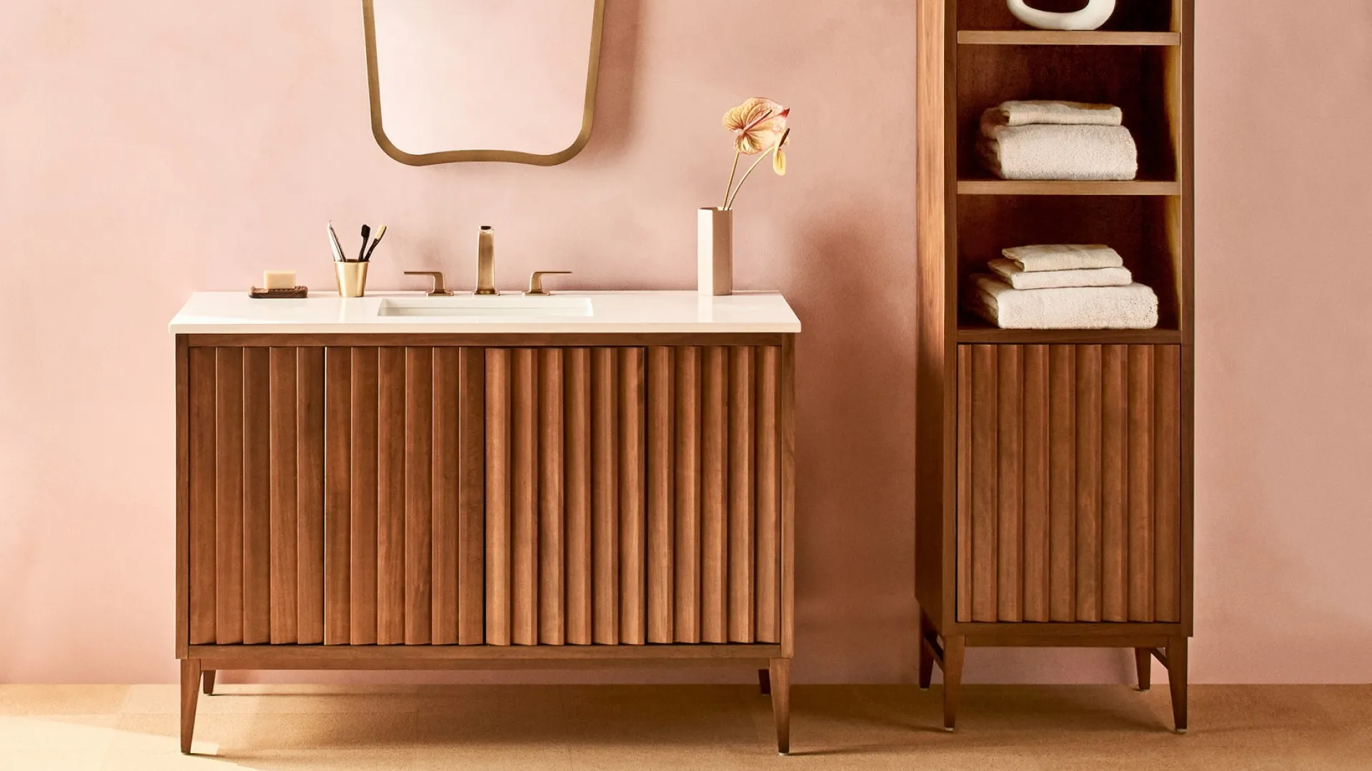 Modern bathroom setup with wooden vanity, unique mirror, tall shelving unit, and soft pink wall background.