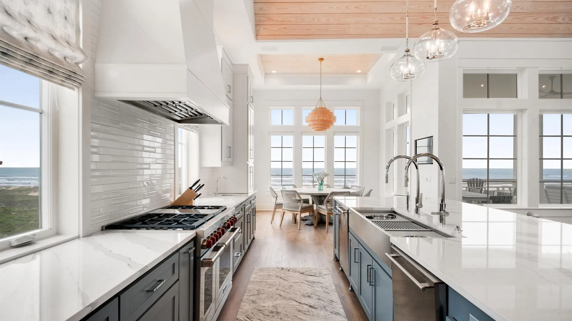 Bright modern kitchen with marble countertops, blue cabinetry, wooden ceiling, and ocean views through large windows.
