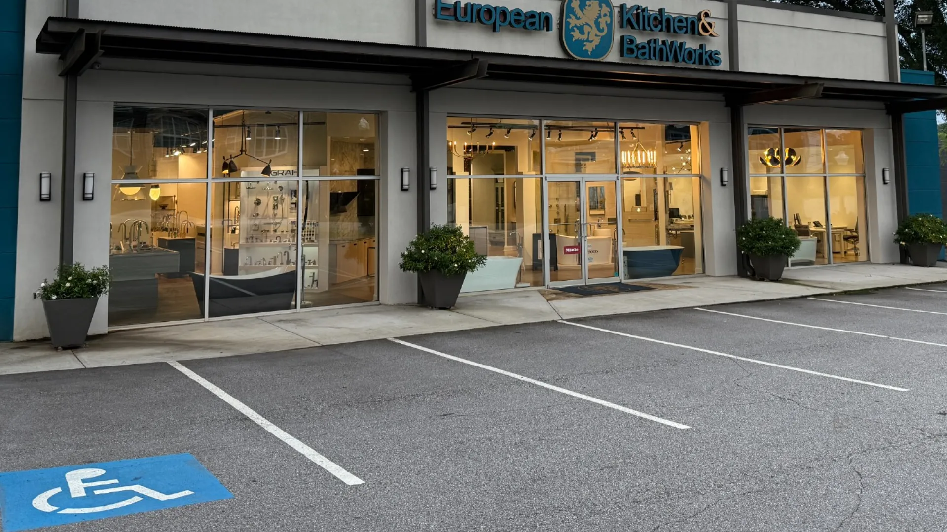 Exterior of European Kitchen & Bath Works showroom with glass windows, potted plants, and empty parking spaces at dusk.
