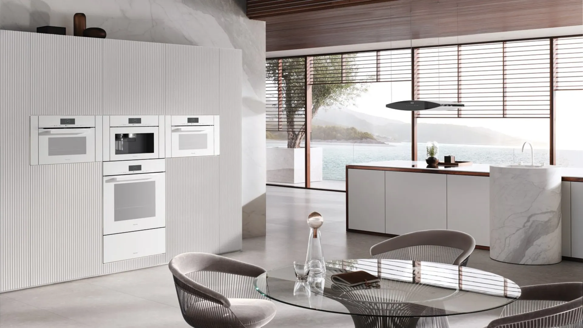 Modern kitchen with built-in white appliances, glass dining table, and large windows overlooking water and mountains.