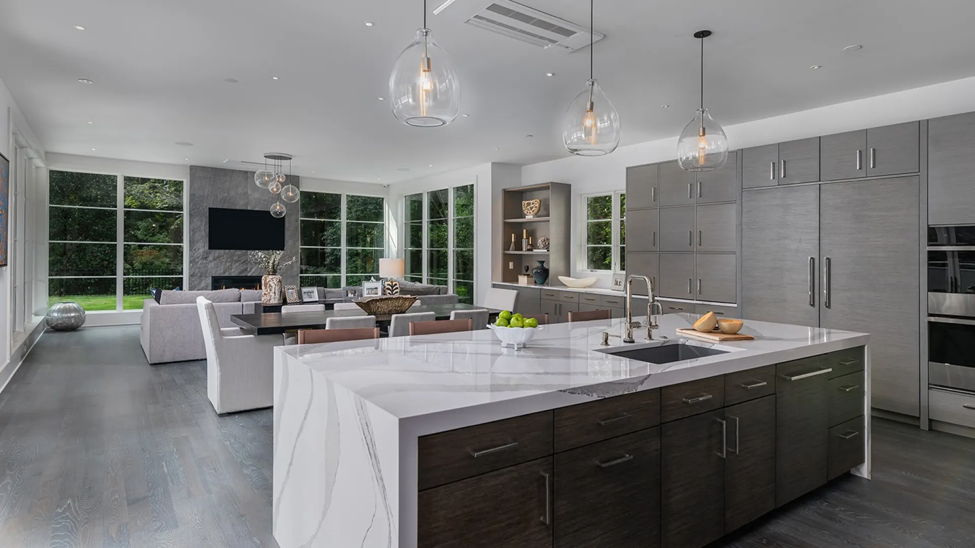 Modern open-concept kitchen and living room with marble island, pendant lights, and large windows overlooking green yard