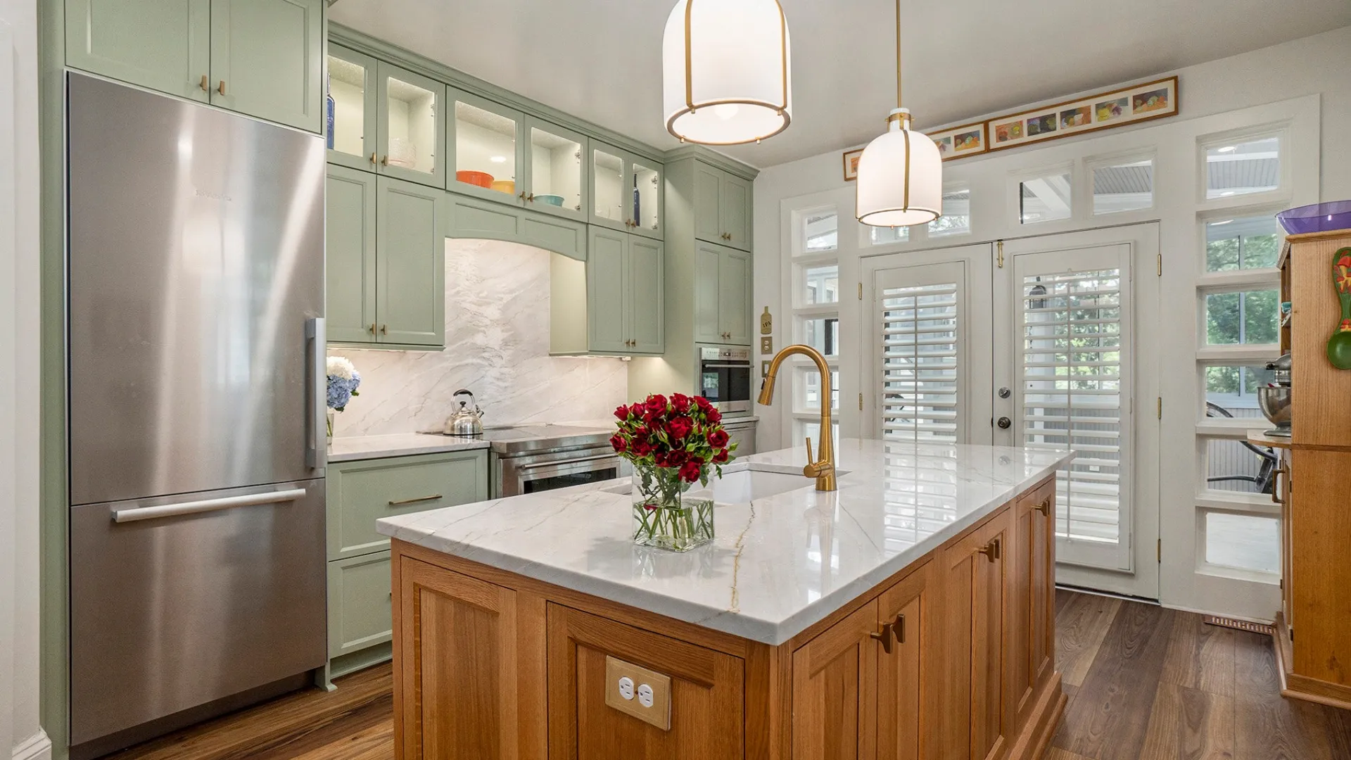 Modern kitchen featuring green cabinets, marble countertops, wooden island, and bright lighting with fresh flowers.