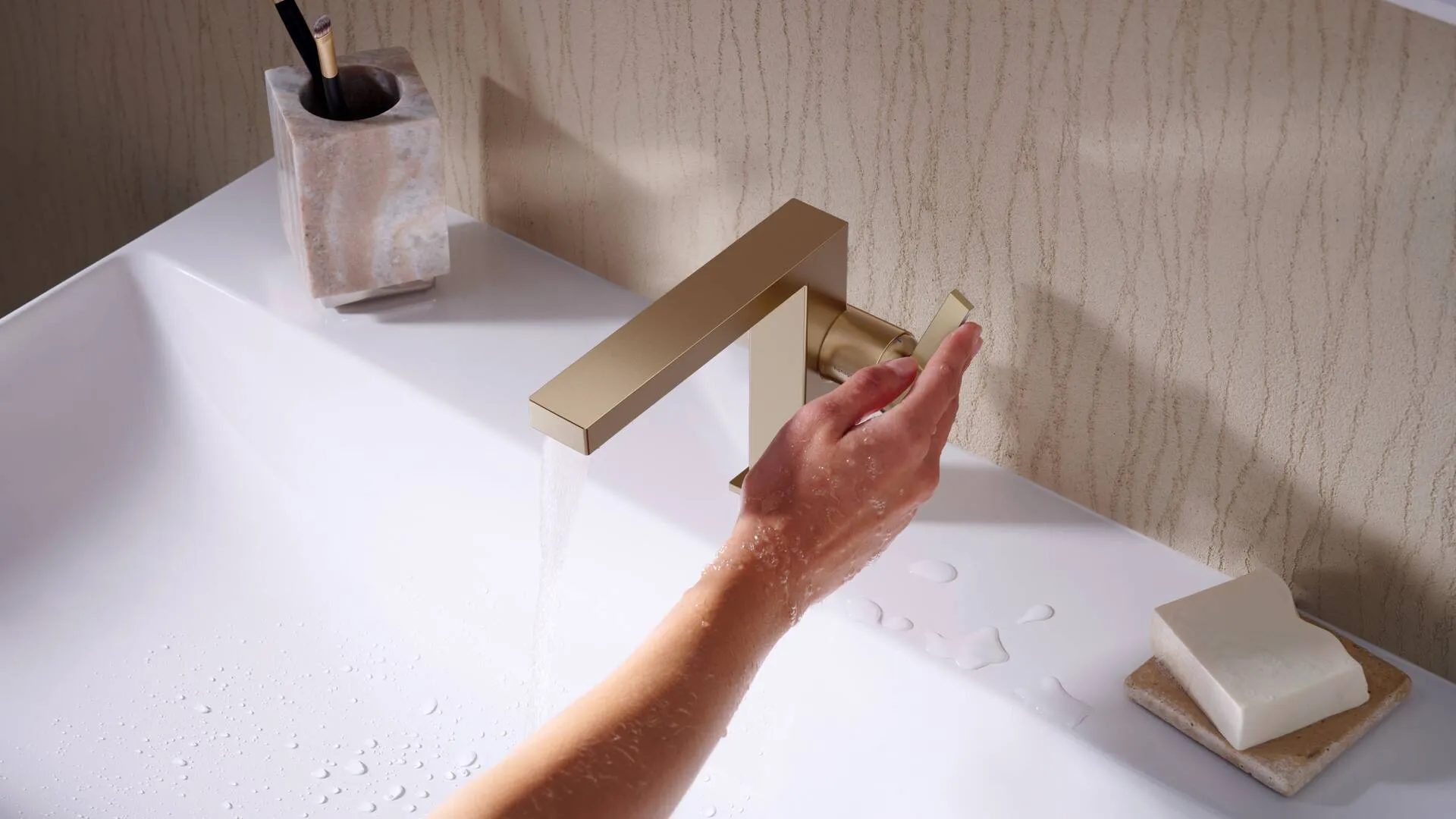 Person washing hands with running water from modern gold faucet in white sink with soap and toothbrush holder.