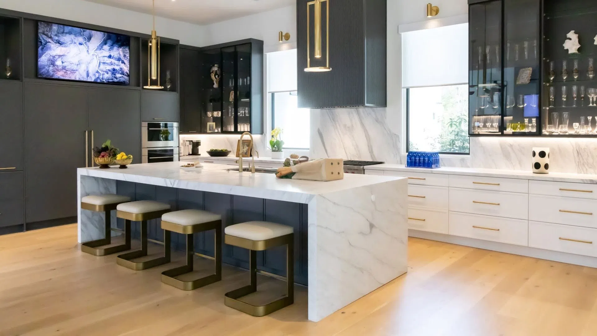 Modern kitchen with marble island, black cabinetry, gold fixtures, and light wooden flooring featuring four stools.