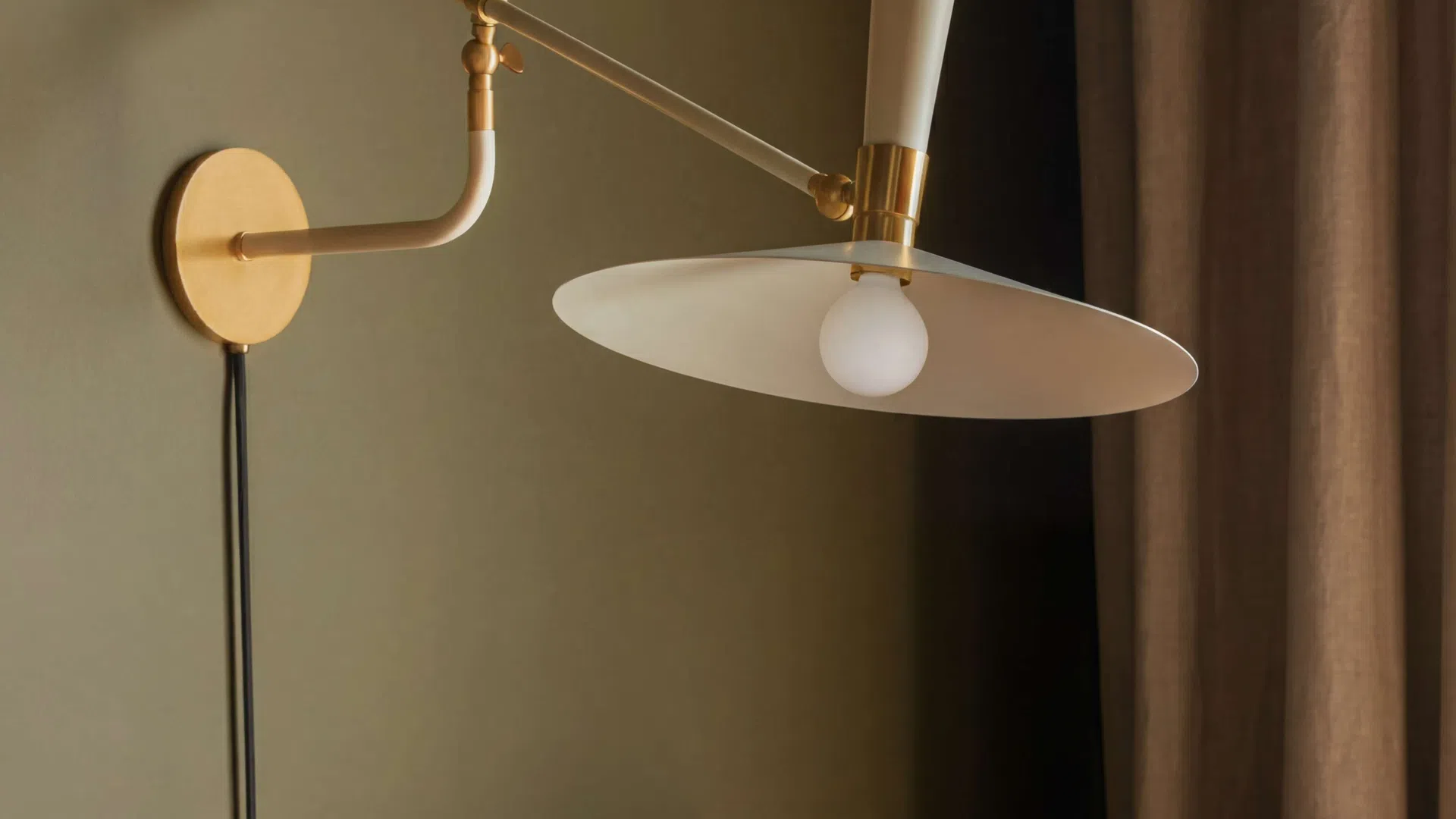 Modern wall-mounted brass lamp with white shade above a patterned velvet sofa corner near a brown curtain.