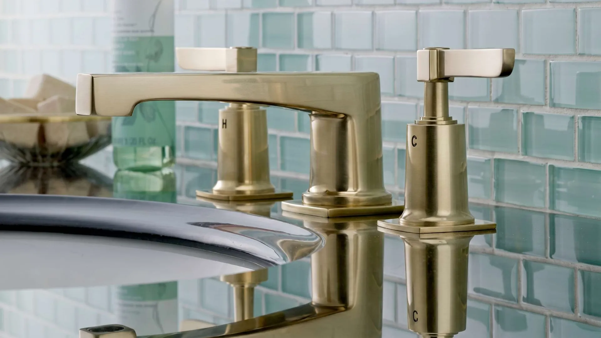 Brushed gold bathroom faucet with hot and cold handles over a sink with tiled backsplash and toiletries