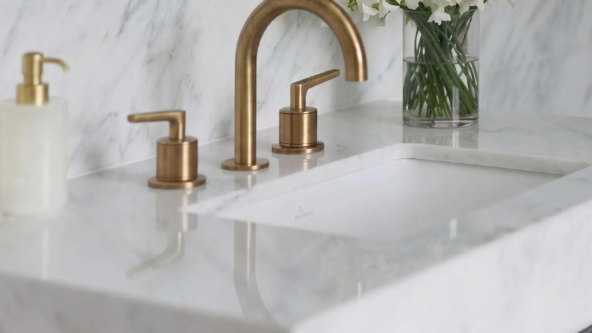 Modern bathroom sink with brass faucet, white marble countertop, and vase of white flowers.