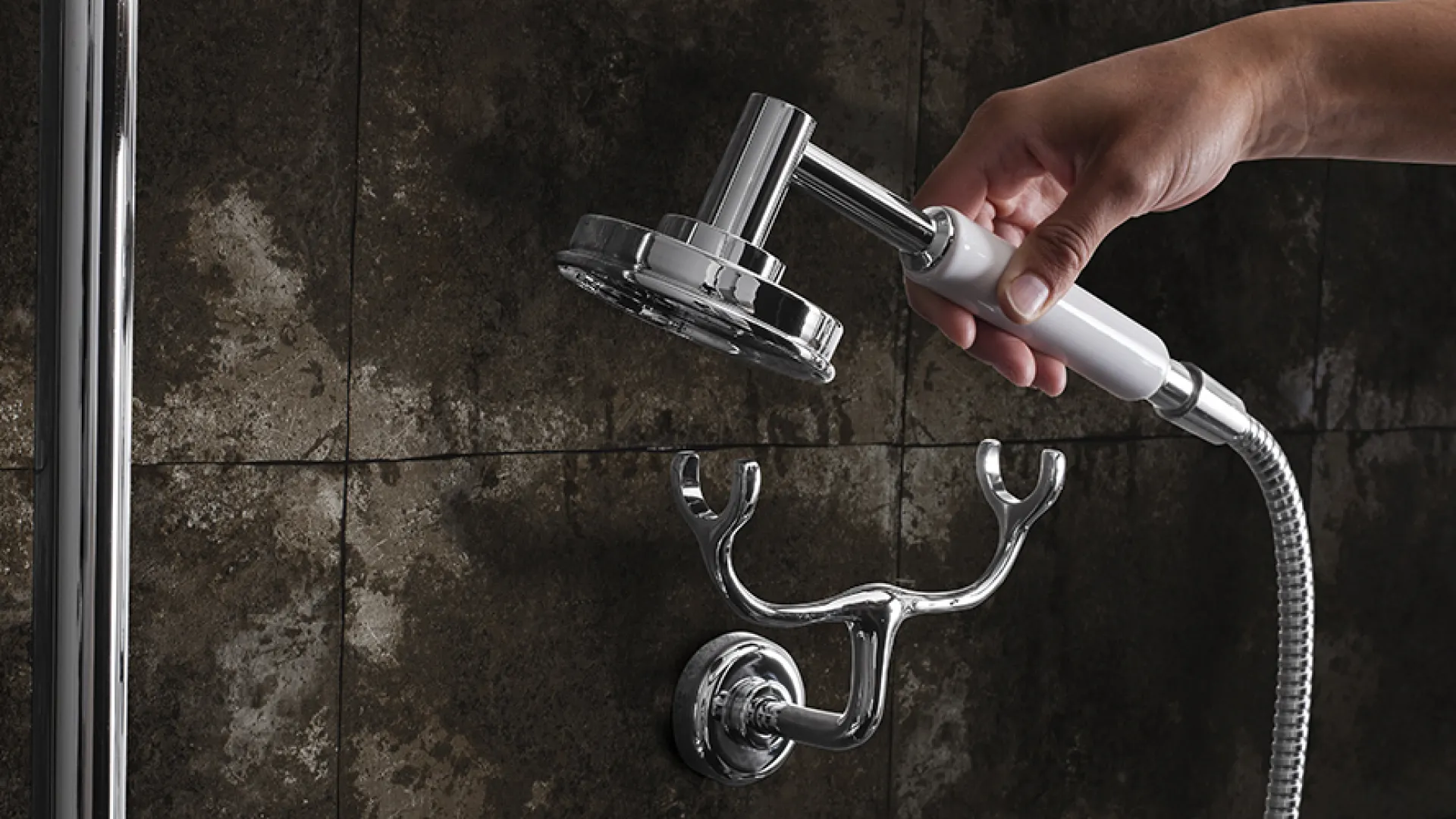 Hand holding a detachable showerhead against dark tiled bathroom wall with chrome fixtures.
