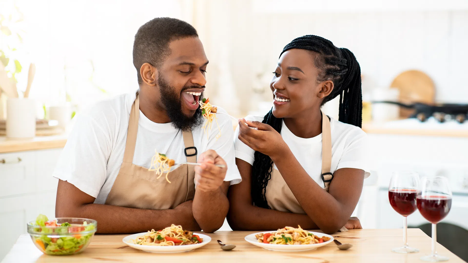 Couple wearing aprons feeding each other spaghetti at a kitchen table with salad and red wine.