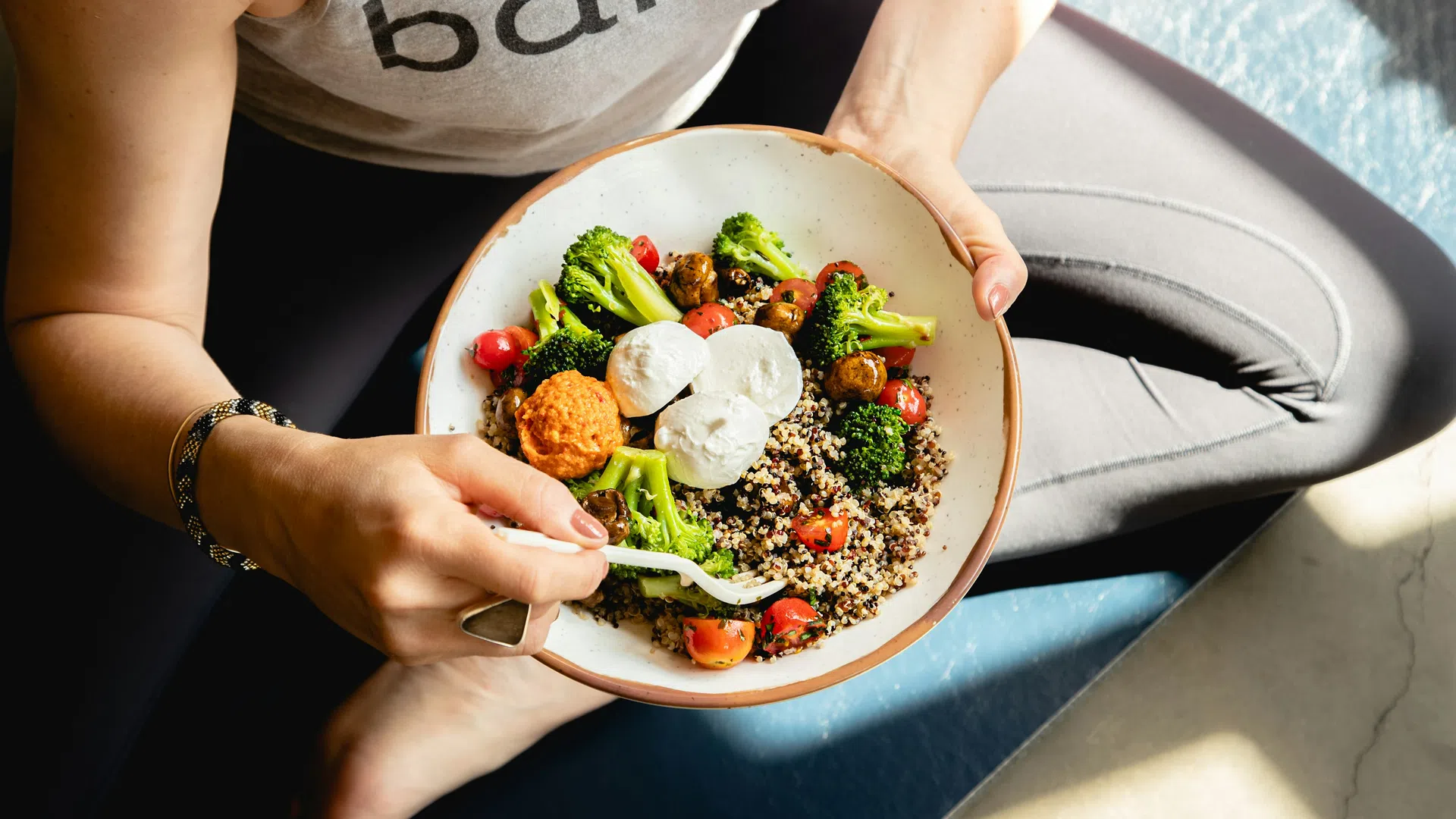 Person holding bowl of quinoa salad with broccoli, cherry tomatoes, falafel, and goat cheese while seated cross-legged.