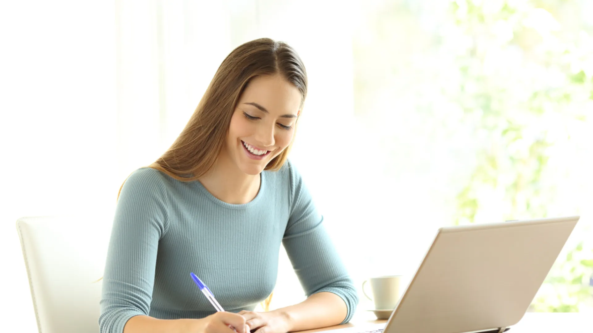 Young woman smiling while writing notes beside an open laptop in a bright workspace