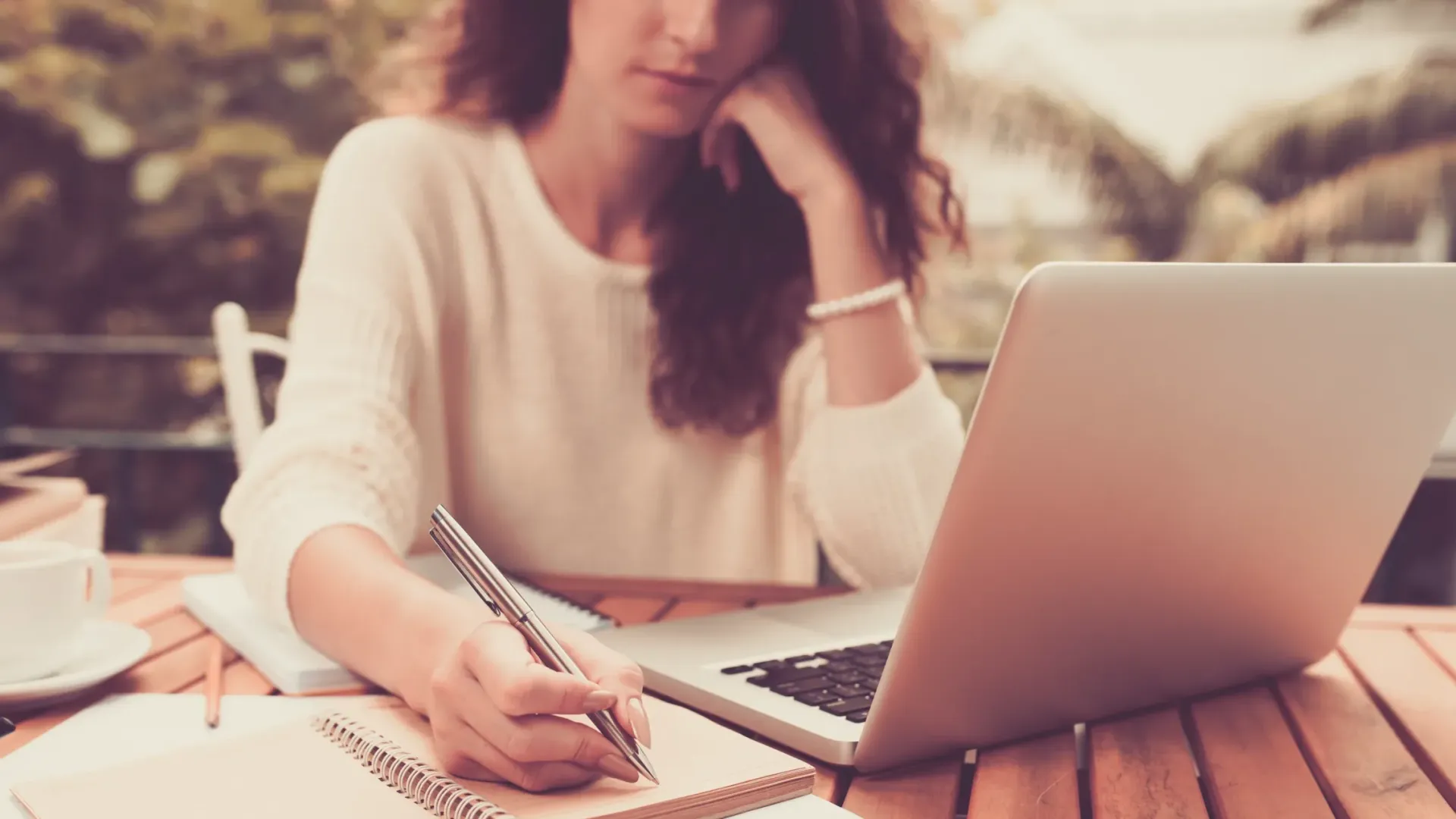 Woman writing notes while working on a laptop at an outdoor wooden table with a coffee cup nearby