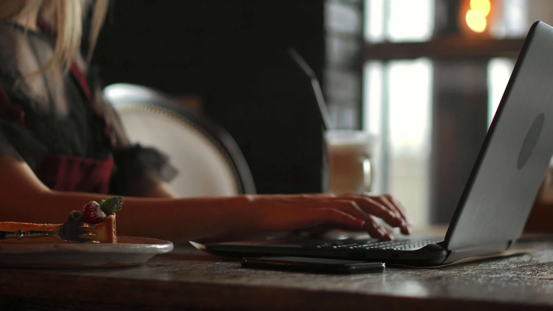 Person typing on a laptop at a wooden table with dessert and coffee in a cozy setting with natural light.