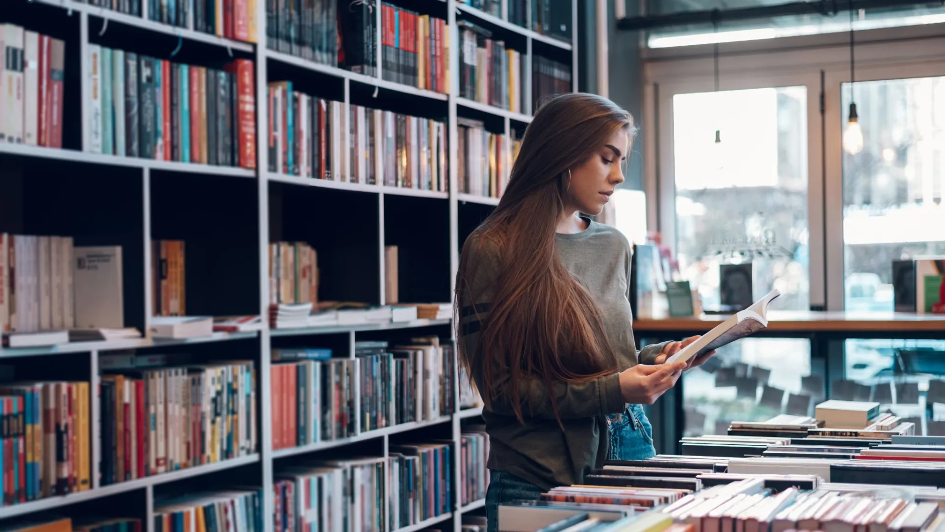 Young woman reading a book in a cozy bookstore filled with shelves and tables of books.