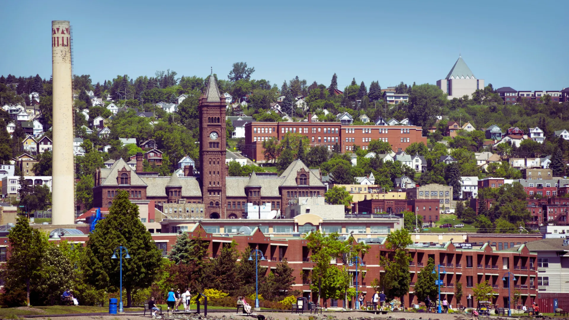 Historic city skyline with old brick buildings, tall smokestack, and green trees under blue sky.