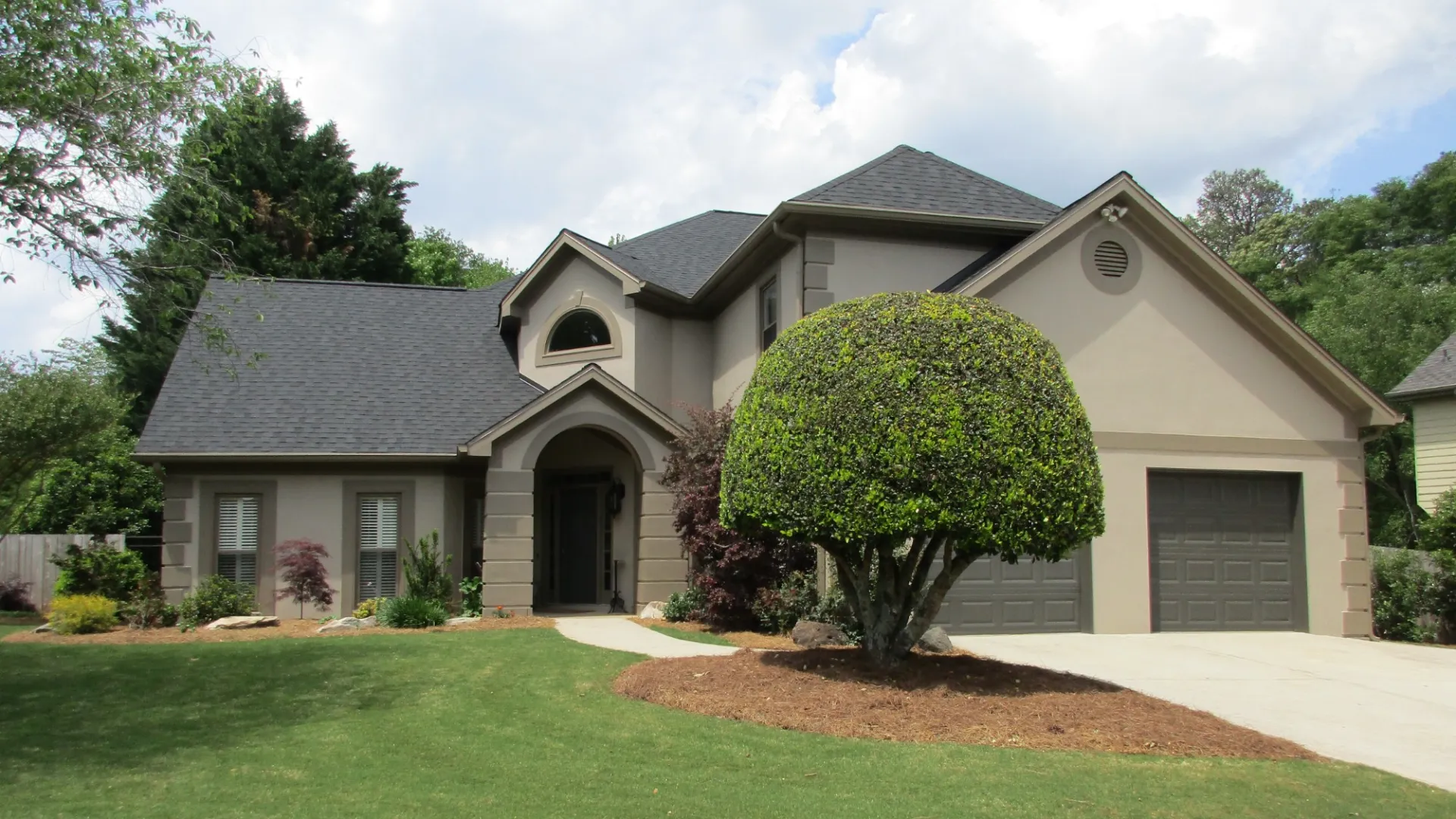 Two-story beige house with dark roof, large manicured tree, and green lawn under partly cloudy sky