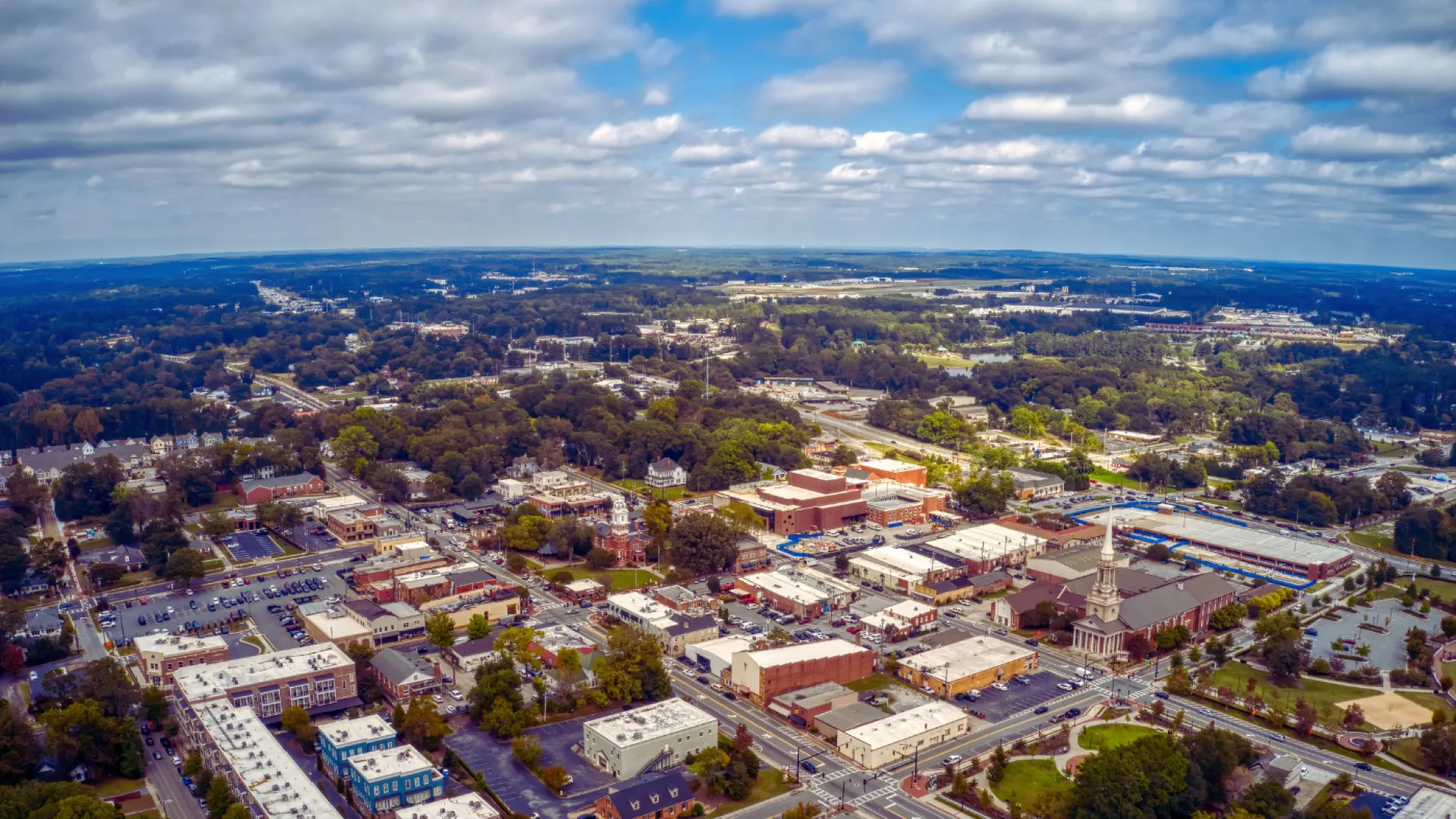 Aerial view of a small town with buildings, a church, streets, and surrounding green trees under a cloudy sky.
