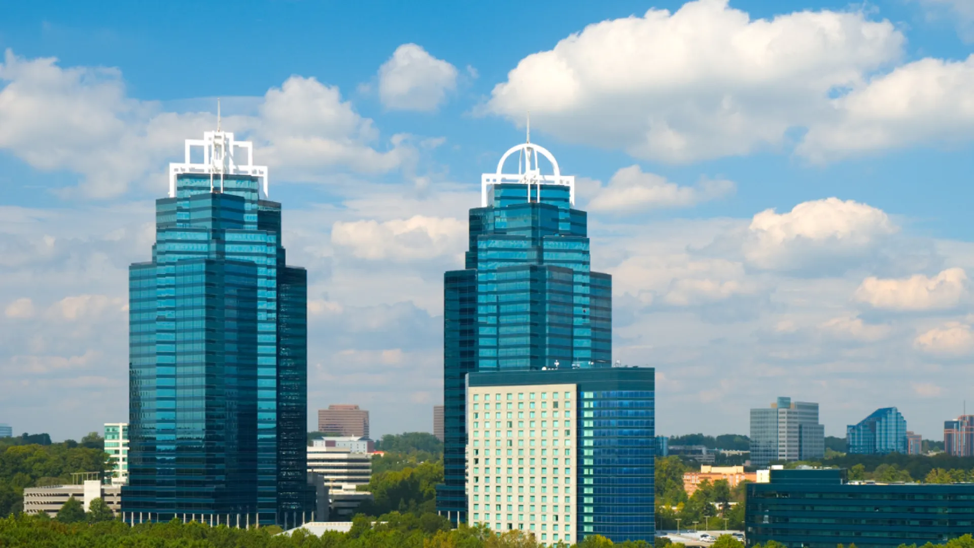 Modern twin glass skyscrapers surrounded by smaller buildings and lush green trees under a partly cloudy sky