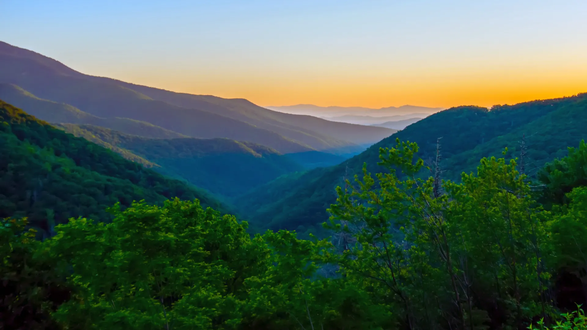 Sunset over layered mountain ranges with green forested hills in the foreground and a clear sky.