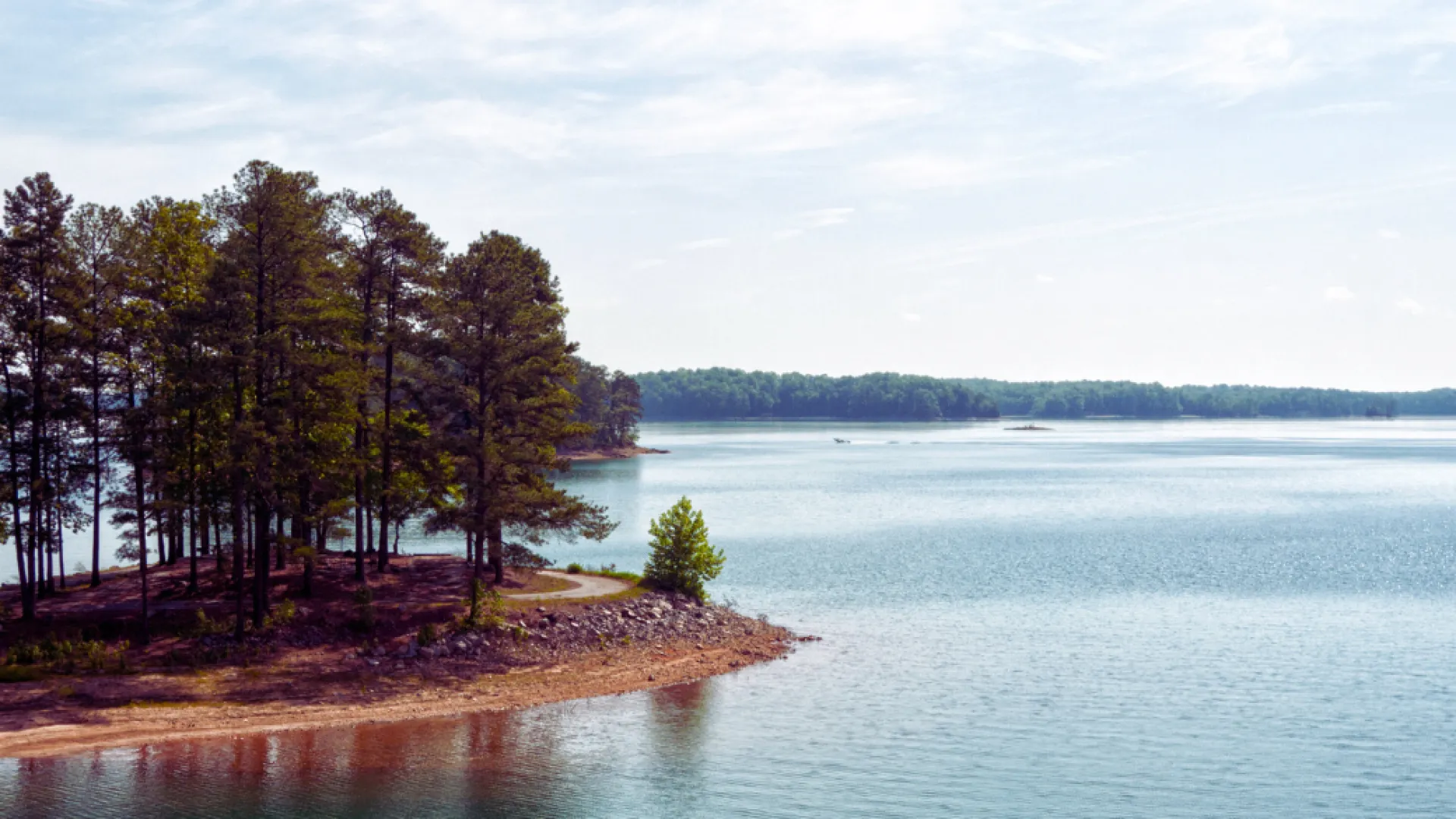 Calm lake with clear blue water and pine trees on a small sandy peninsula under a partly cloudy sky.