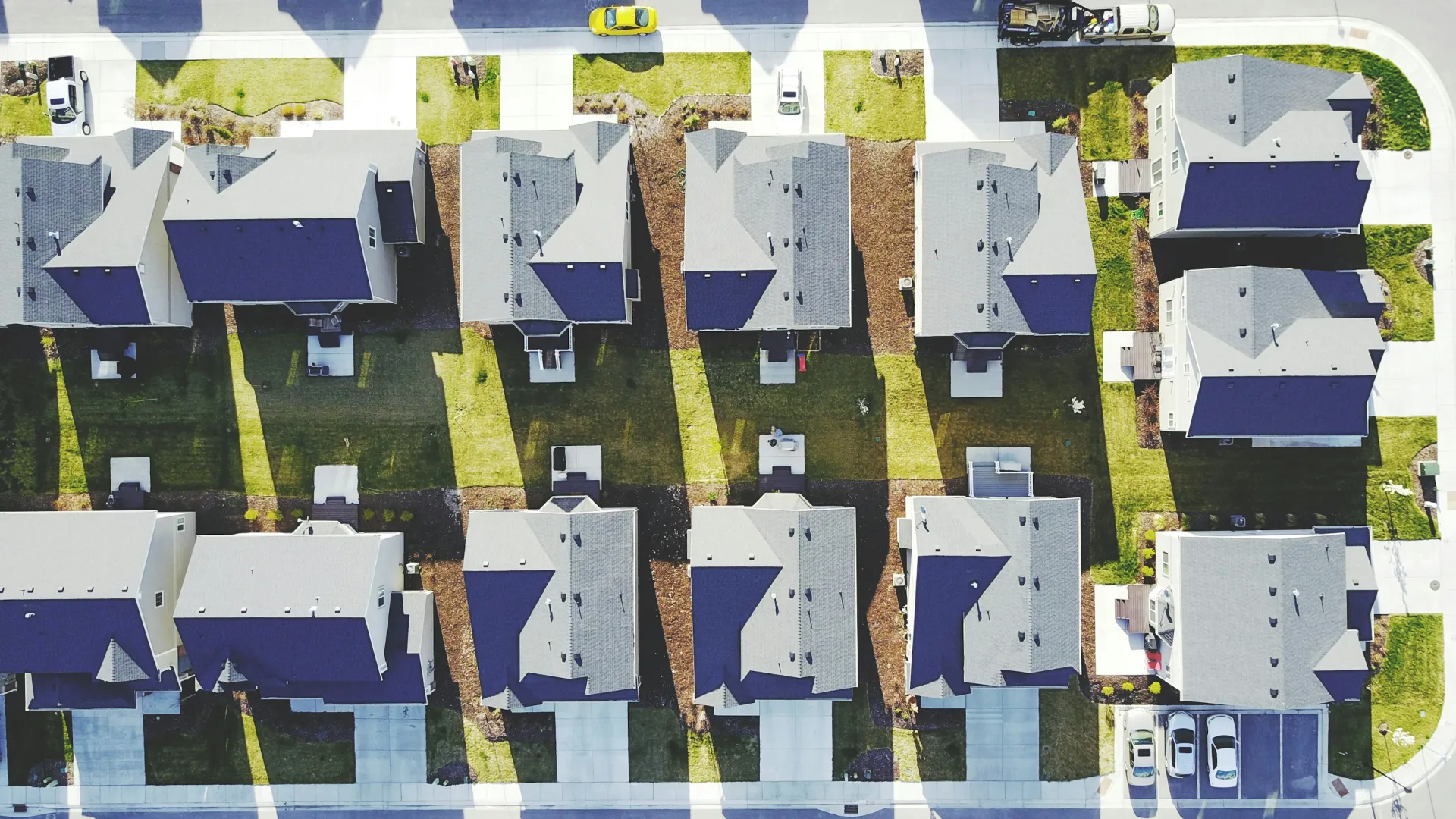Aerial view of a suburban neighborhood showing rows of houses, driveways, lawns, and parked cars on sunny day.