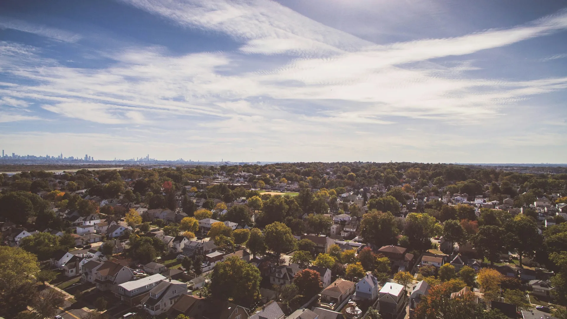 Aerial view of a suburban neighborhood with houses, trees, and a city skyline in the distance under a partly cloudy sky
