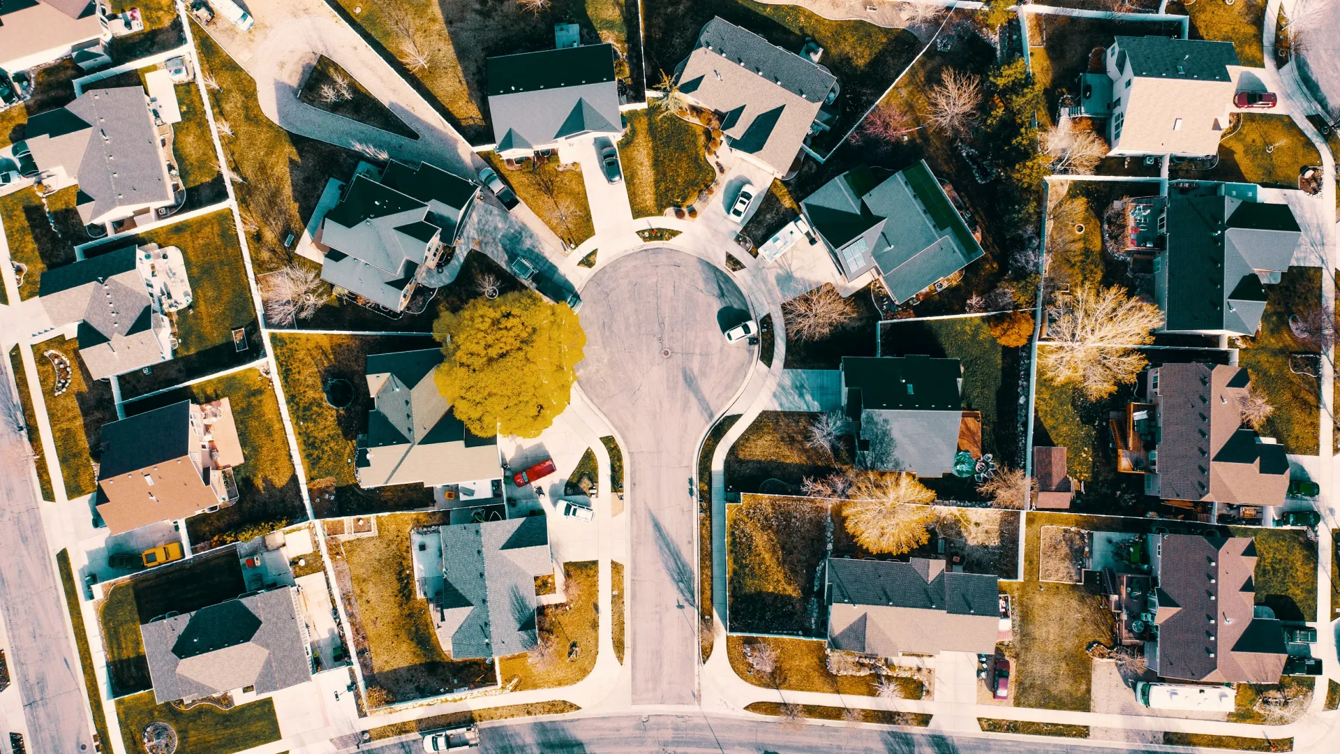 Aerial view of a suburban cul-de-sac with houses, driveways, cars, and trees casting shadows on lawns.