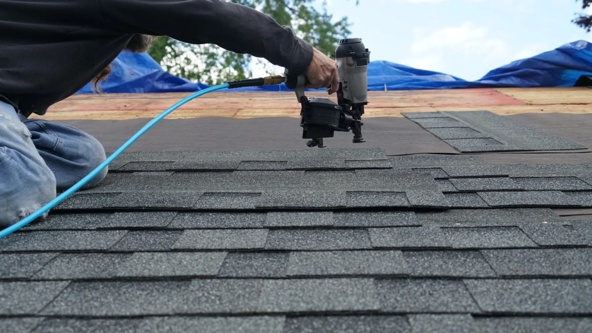 Worker using nail gun to install asphalt shingles on a roof under a blue sky.