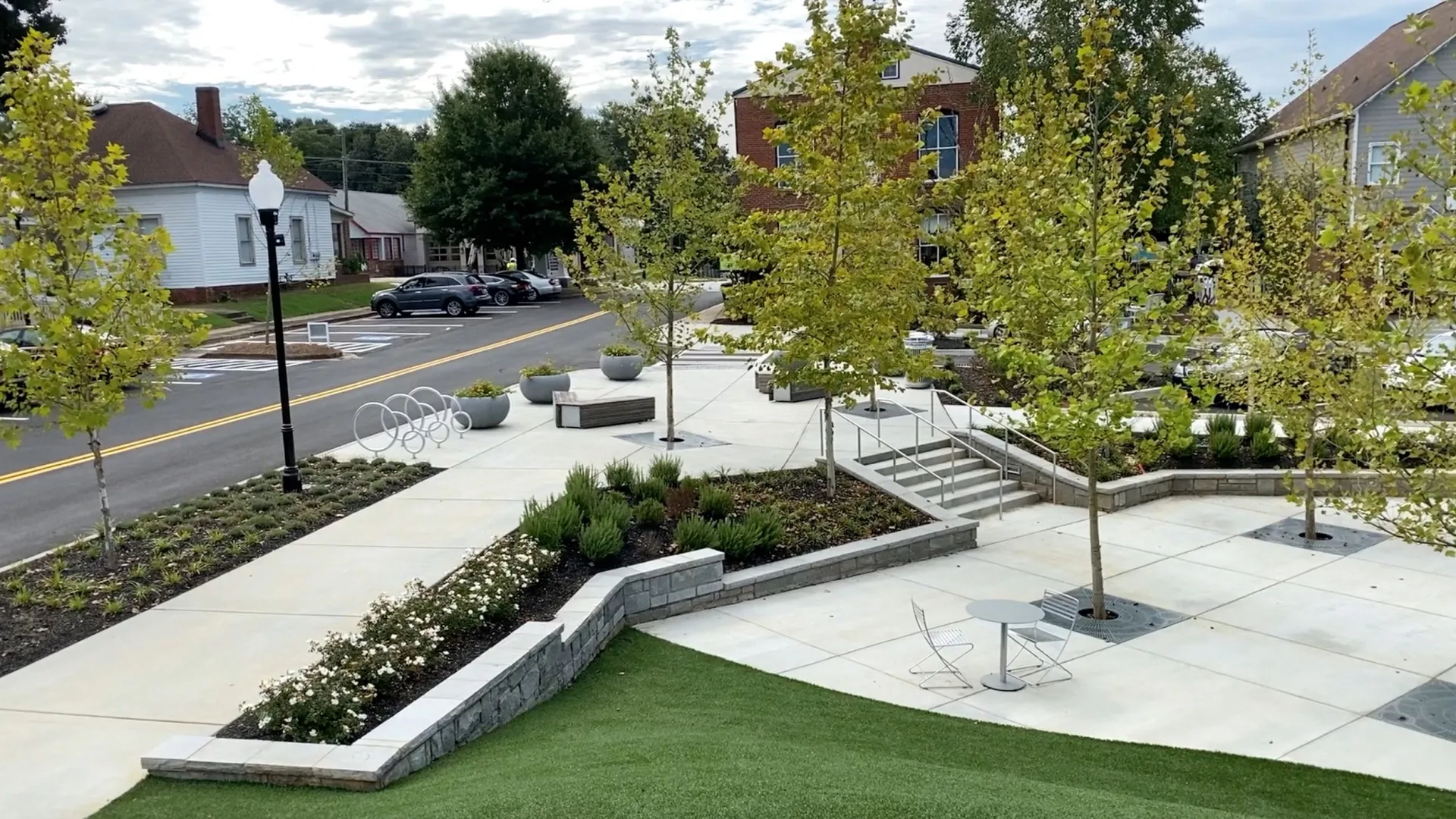 Modern urban park with concrete paths, trees, benches, greenery, and a view of surrounding buildings and street.