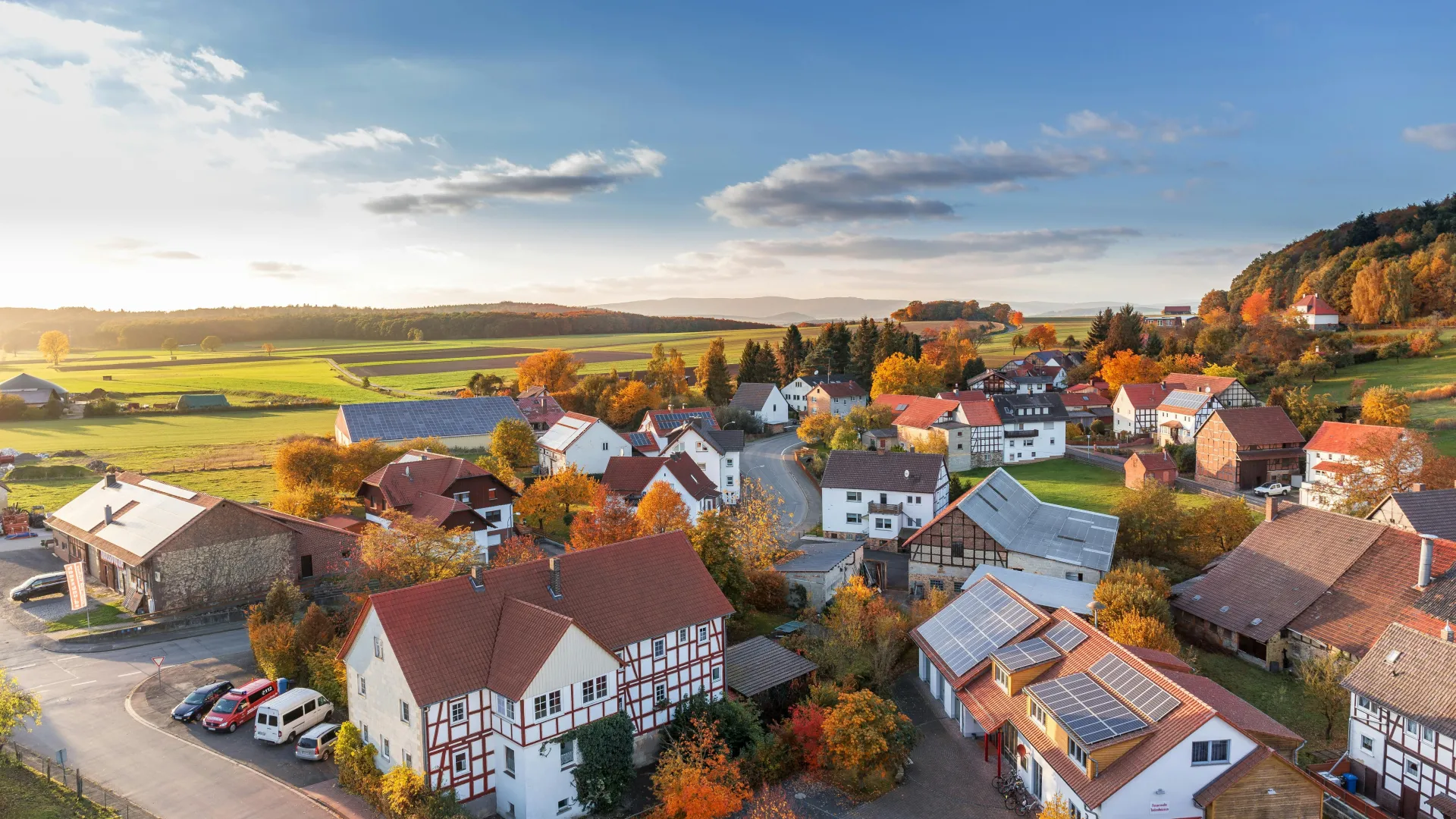 Aerial view of a quaint village with traditional houses, autumn foliage, and green fields under a partly cloudy sky.